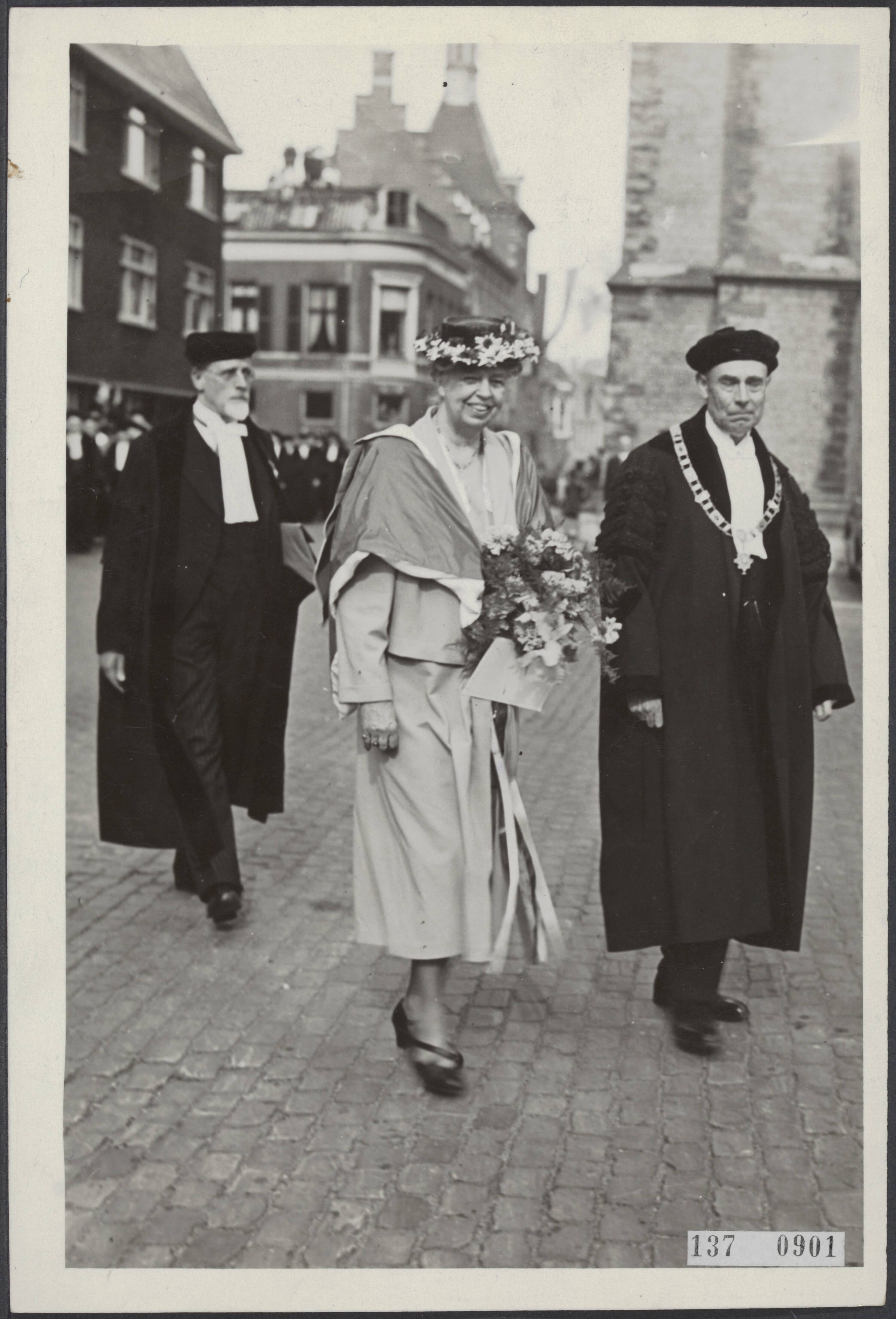 Actualiteiten, binnenland — In tegenwoordigheid van koningin Wilhelmina, prinses Juliana en prins Bernhard werd aan Eleanor Roosevelt het eredoctoraat in de rechtsgeleerdheid van de Rijksuniversiteit te Utrecht aangeboden. De plechtigheid vond plats in de Domkerk te Utrecht. Mevr. Roosevelt begeeft zich na de plechtigheid, in het bezit van de bul, van de Domkerk terug naar de universiteit; naast haar de rector-magnificus, prof. U.G. Bijlsma, achter haar de promotor prof. dr. S. van Brakel — Foto: Fotograaf Onbekend / Anefo. Licentie: Set B: Rechtenvrij / Publiek Domein. Bron: Nationaal Archief. http://hdl.handle.net/10648/af6a97b4-d0b4-102d-bcf8-003048976d84