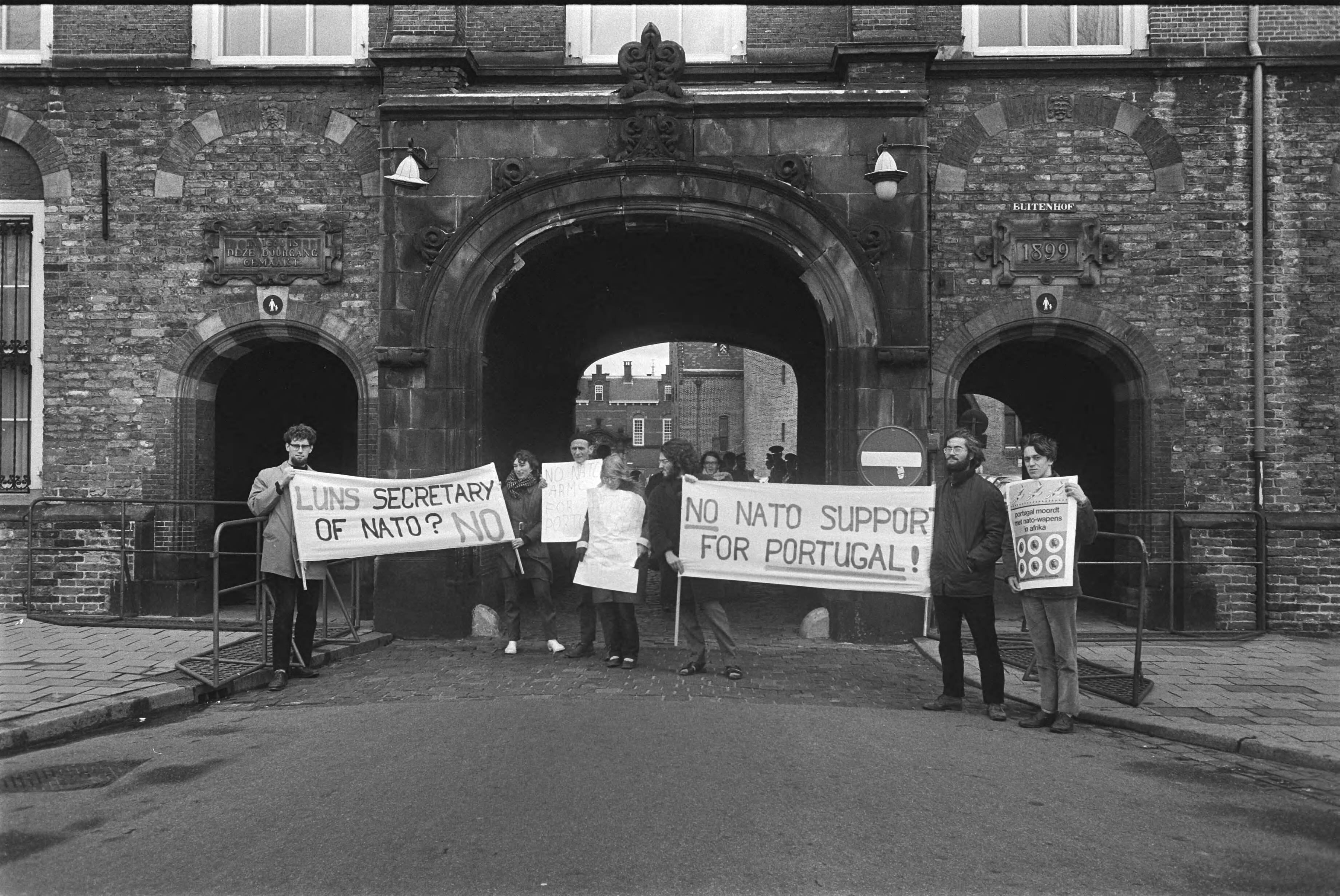 Fotocollectie Anefo — Demonstranten met borden tegen NATO-vergadering voor toegangspoort Binnenhof Den Haag — Foto: Koch, Eric / Anefo. Licentie: Set B: Rechtenvrij / Publiek Domein. Bron: Nationaal Archief. http://hdl.handle.net/10648/aba977ee-d0b4-102d-bcf8-003048976d84