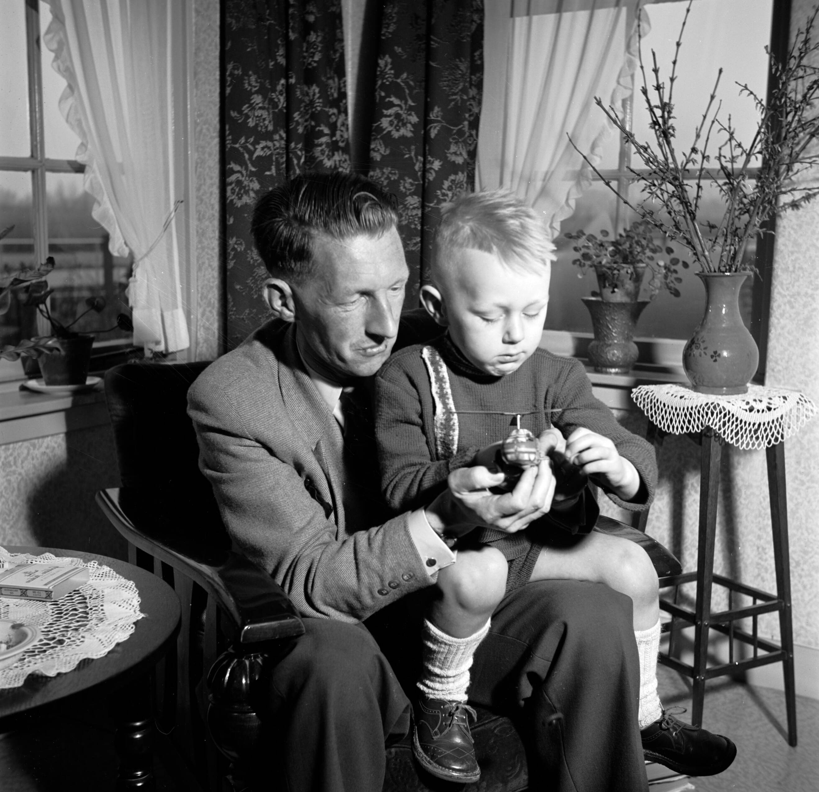 Fotocollectie Van de Poll — Gerard met zijn vierjarig zoontje Gerrie op schoot met een speelgoed helikopter. Links en bloementafeltje met een vaas met uitbottende takken — Foto: Poll, Willem van de. Licentie: Set B: Rechtenvrij / Publiek Domein. Bron: Nationaal Archief. http://hdl.handle.net/10648/aeccd164-d0b4-102d-bcf8-003048976d84