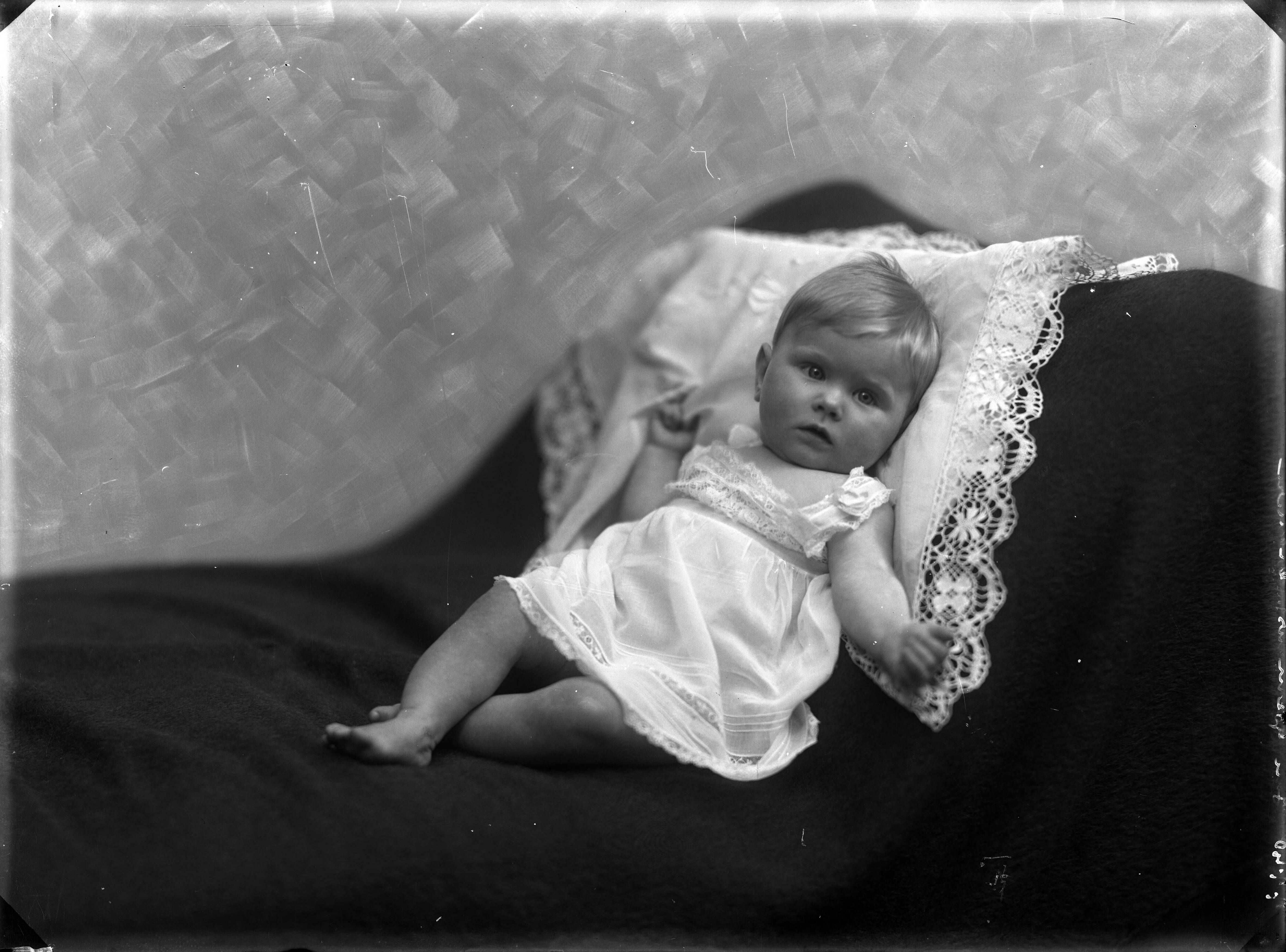 Berg, van den (BERG 050) — Studio-opnames. Babymeisje in een wit jurkje op een wit kanten kussen tegen de leuning van een bank. Haarlem, Nederland. 1918-1930. 7740 Jan Goemans — Foto: Berg, Th.C.J. van den / Spaarnestad Photo (05887). Licentie: Set B: Rechtenvrij / Publiek Domein. Bron: Nationaal Archief. http://hdl.handle.net/10648/35429da7-c773-a382-e063-6df0900a6686
