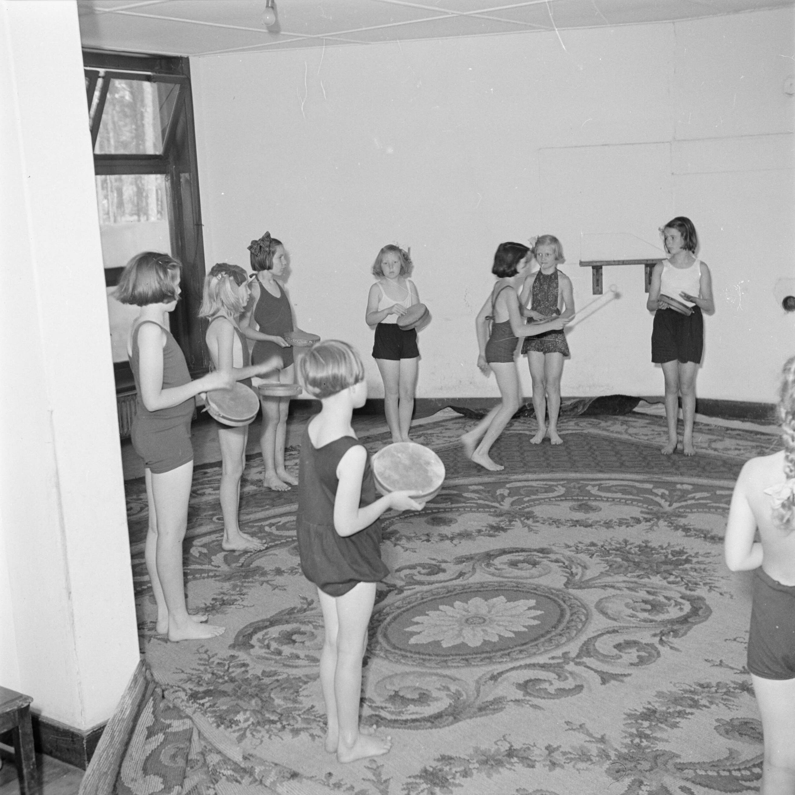Prinses Beatrix en prinses Irene op de Werkplaats Kindergemeenschap van Kees Boeke te Bilthoven — Prinses Beatrix en andere leerlingen met tamboerijnen in hun hand tijdens de gymnastiekles — Foto: Poll, Willem van de. Licentie: Set B: Rechtenvrij / Publiek Domein. Bron: Nationaal Archief. http://hdl.handle.net/10648/aee8162c-d0b4-102d-bcf8-003048976d84