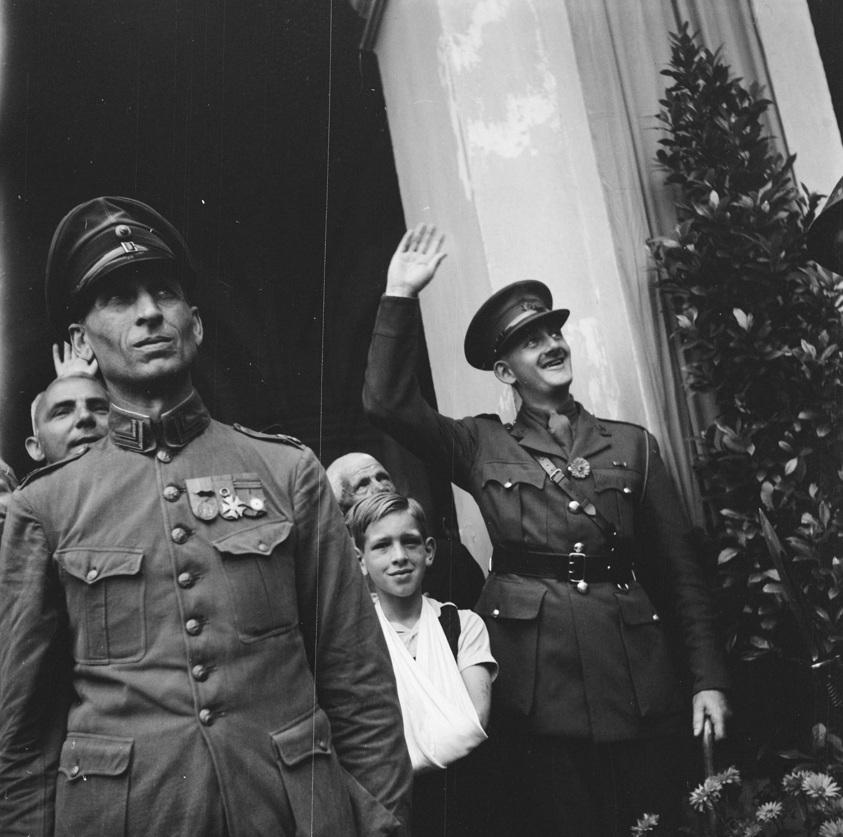 Koningin Wilhelmina terug in Den Haag — Links een KNIL-militair en rechts een Canadese? officier die zwaait. Tussen hen in een jongen met een mitella — Foto: Meijboom, Marius / Anefo. Licentie: Set B: Rechtenvrij / Publiek Domein. Bron: Nationaal Archief. http://hdl.handle.net/10648/ac0f055a-d0b4-102d-bcf8-003048976d84