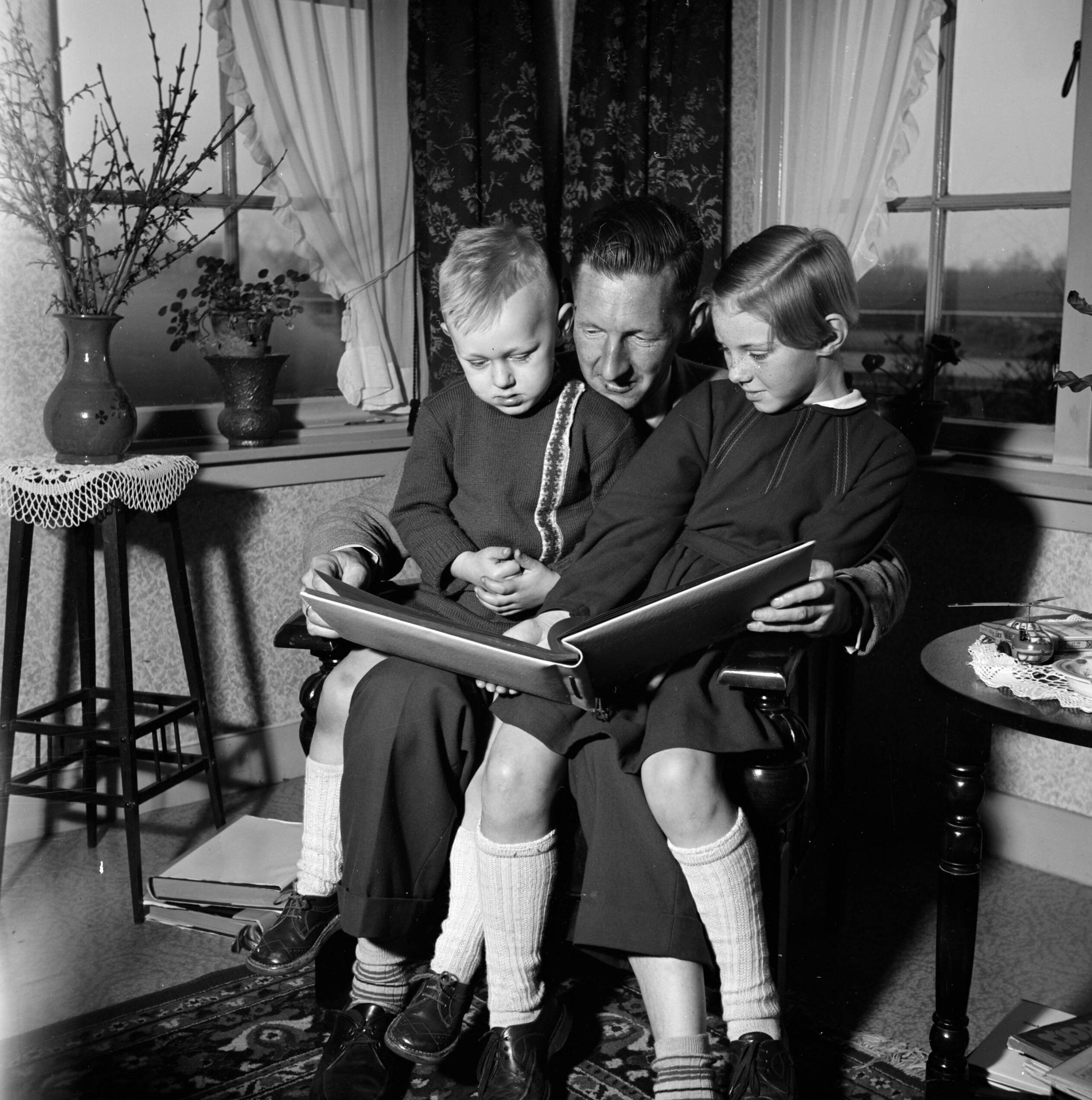 Fotocollectie Van de Poll — Gerard met zijn beide kinderen op schoot terwijl ze een fotoalbum bekijken. Links een bloementafeltje met een vaas met uitbottende takken — Foto: Poll, Willem van de. Licentie: Set B: Rechtenvrij / Publiek Domein. Bron: Nationaal Archief. http://hdl.handle.net/10648/aeccd3bc-d0b4-102d-bcf8-003048976d84