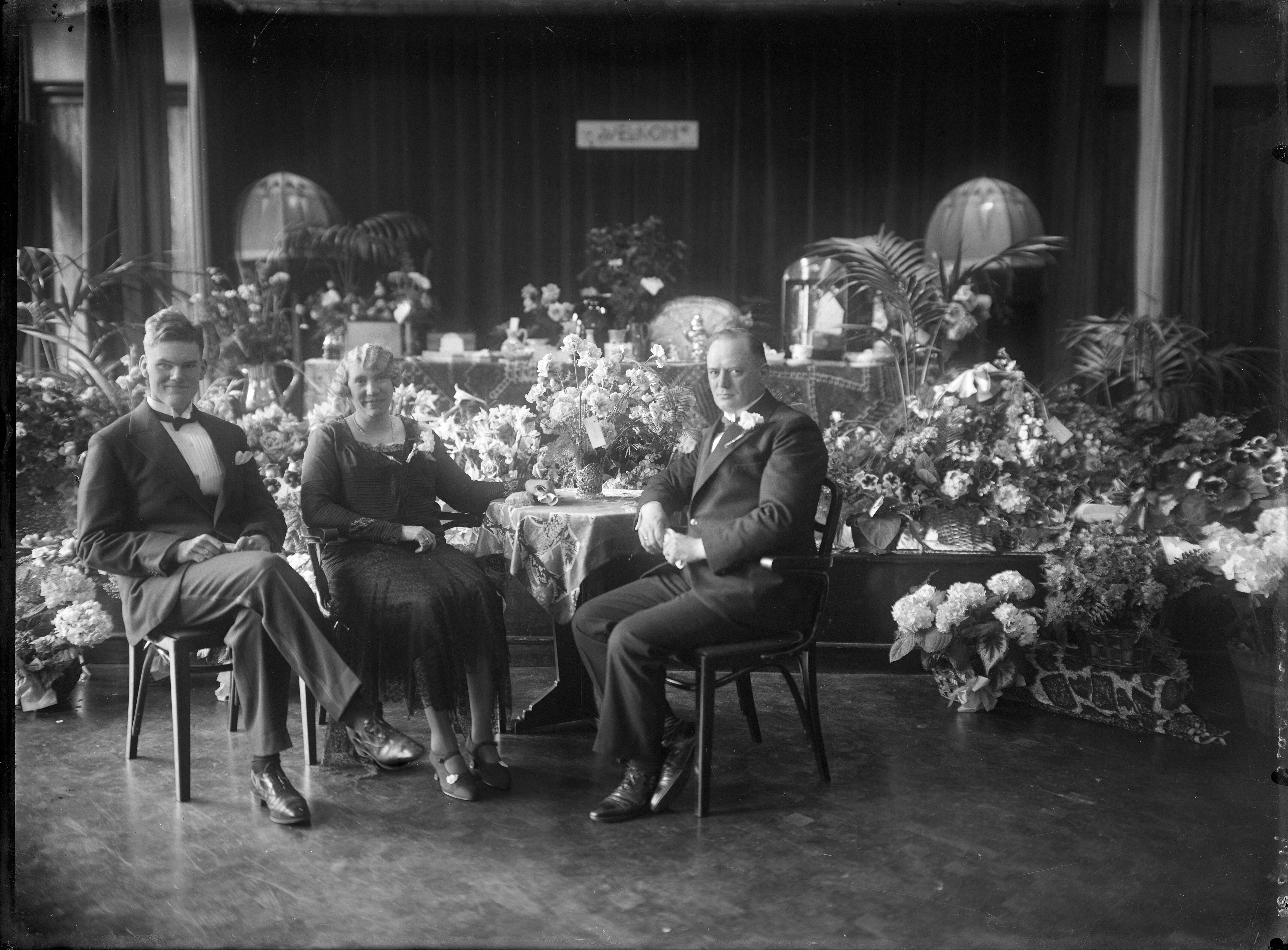 Berg, van den (BERG 050) — Feesten. Een jubilerend echtpaar met hun zoon in een met heel veel bloemen versierde feestlokatie. Nederland. 1918-1930.8519 Braakman (niet goed leesbaar) — Foto: Berg, Th.C.J. van den / Spaarnestad Photo (05887). Licentie: Set B: Rechtenvrij / Publiek Domein. Bron: Nationaal Archief. http://hdl.handle.net/10648/35429da7-c802-a382-e063-6df0900a6686