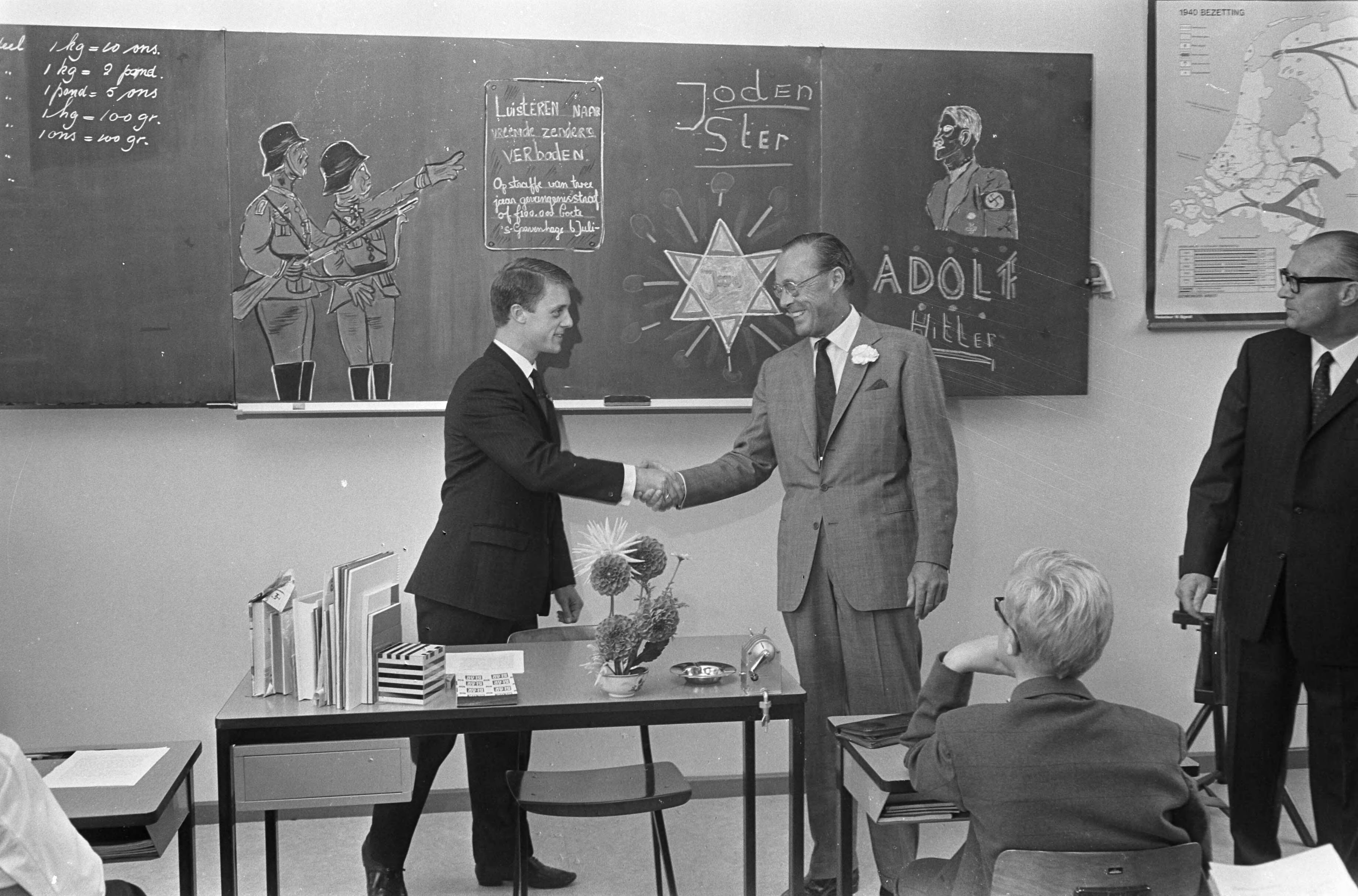 Prins Bernhard opent de Prins Bernhard school te Leeuwarden — Prins Bernhard staat in een klas bij bordtekeningen over de bezetting — Foto: Fotograaf Onbekend / Anefo. Licentie: Set B: Rechtenvrij / Publiek Domein. Bron: Nationaal Archief. http://hdl.handle.net/10648/ab4c43e4-d0b4-102d-bcf8-003048976d84