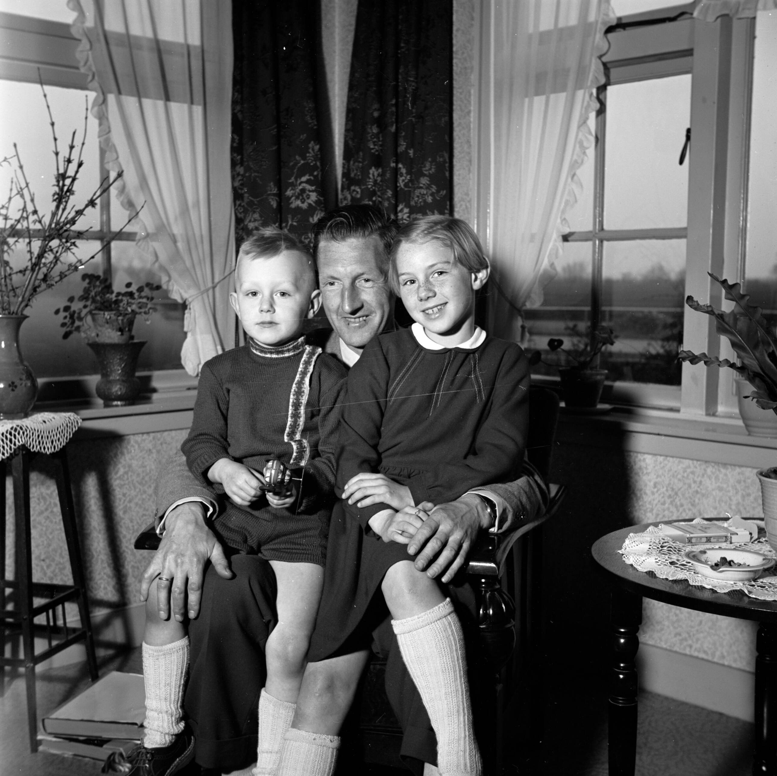 Fotocollectie Van de Poll — Gerard met zijn beide kinderen op schoot, terwijl ze een fotoalbum bekijken. Links een bloementafeltje met een vaas met uitbottende takken — Foto: Poll, Willem van de. Licentie: Set B: Rechtenvrij / Publiek Domein. Bron: Nationaal Archief. http://hdl.handle.net/10648/aeccd47a-d0b4-102d-bcf8-003048976d84