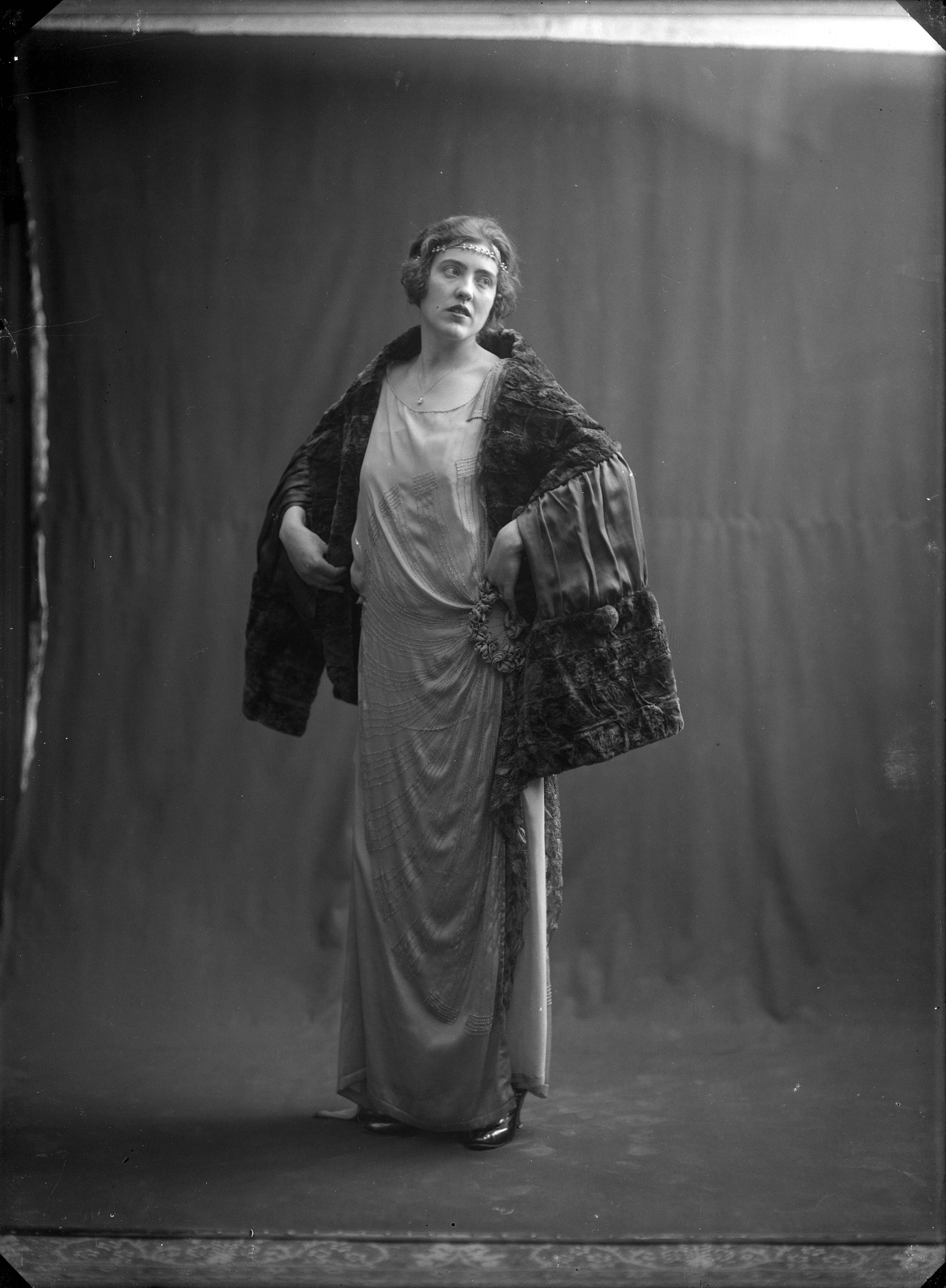 Mode, beauty — Studio-opnames. Een dame in een lange jurk met drapering op de heuo bijeengehouden door ronde versiering. Om de schouders een bontstola met pellerinemouwen. Zonder naam. Haarlem, Nederland 1918-1930. — Foto: Berg, Th.C.J. van den / Spaarnestad Photo (05887). Licentie: Set B: Rechtenvrij / Publiek Domein. Bron: Nationaal Archief. http://hdl.handle.net/10648/35429da7-c55b-a382-e063-6df0900a6686