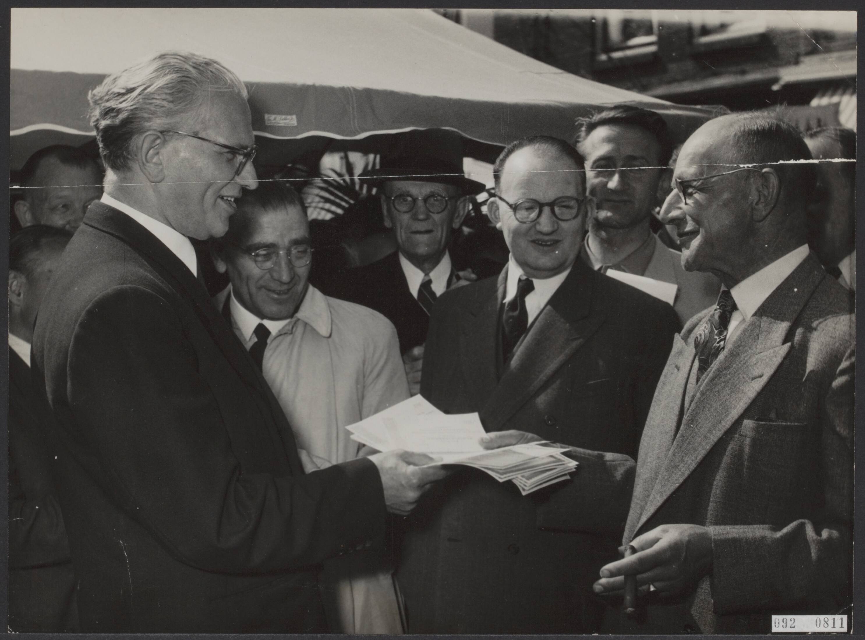 Fotocollectie Elsevier Binnenland — Minister In t Veld verkoopt spaarbrieven Zaandam. Rechts op de foto Dhr. Plooijer — Foto: Noske, J.D. / Anefo. Licentie: Set B: Rechtenvrij / Publiek Domein. Bron: Nationaal Archief. http://hdl.handle.net/10648/ae252996-d0b4-102d-bcf8-003048976d84