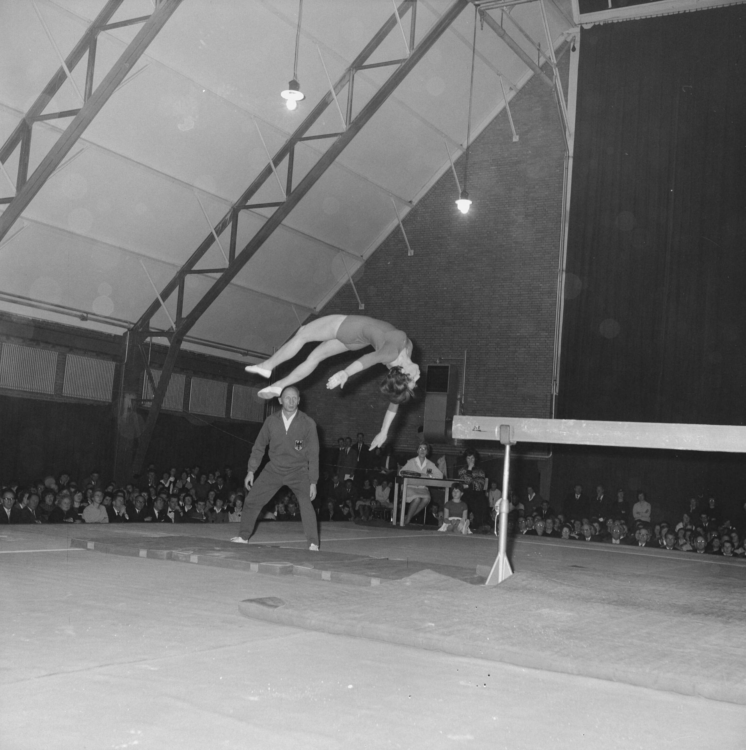 Fotocollectie Anefo — Turninterland Nederland-West-Duitsland in de Centrale Veilinghal te Aalsmeer. Ursel Schepan (Duitsland) bij afsprong evenwichtsbalk. In de achtergrond trainer Niko Szabo — Foto: Nijs, Jac. de / Anefo. Licentie: Set B: Rechtenvrij / Publiek Domein. Bron: Nationaal Archief. http://hdl.handle.net/10648/aa20032a-d0b4-102d-bcf8-003048976d84