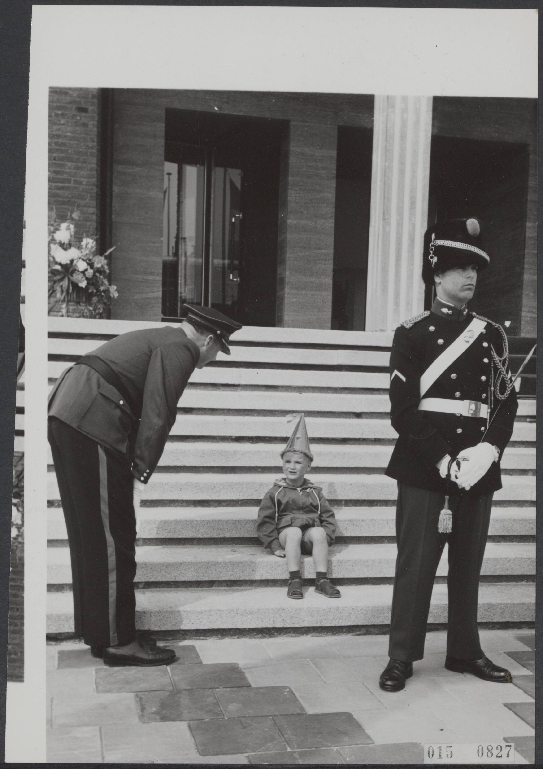 Fotocollectie Elsevier Binnenland — Een kleine jongen met feestmuts zit op de trappen van het nieuwe stadhuis van Vlissingen, dat geopend wordt in het bijzijn van koningin Juliana. Een adjudant van politie praat met de jongen — Foto: Nijs, Jac. de / Anefo. Licentie: Set B: Rechtenvrij / Publiek Domein. Bron: Nationaal Archief. http://hdl.handle.net/10648/adb64760-d0b4-102d-bcf8-003048976d84