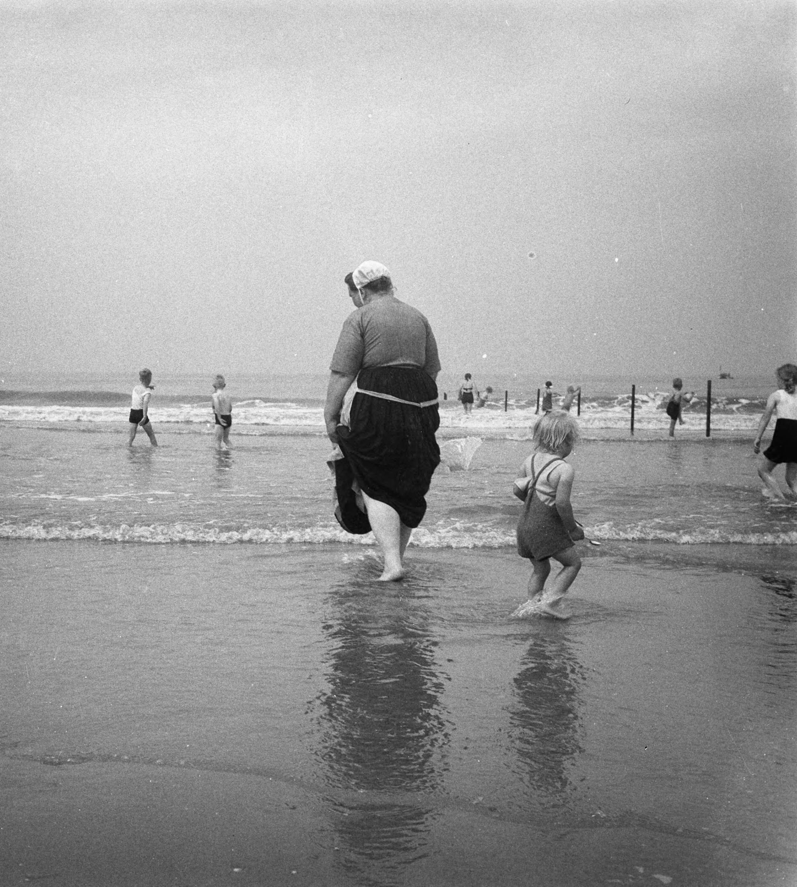 Fotocollectie Anefo — Scheveningse vissersvrouw met kind aan 't pootje baden — Foto: Pot, Harry / Anefo. Licentie: Set B: Rechtenvrij / Publiek Domein. Bron: Nationaal Archief. http://hdl.handle.net/10648/a8ede3e6-d0b4-102d-bcf8-003048976d84
