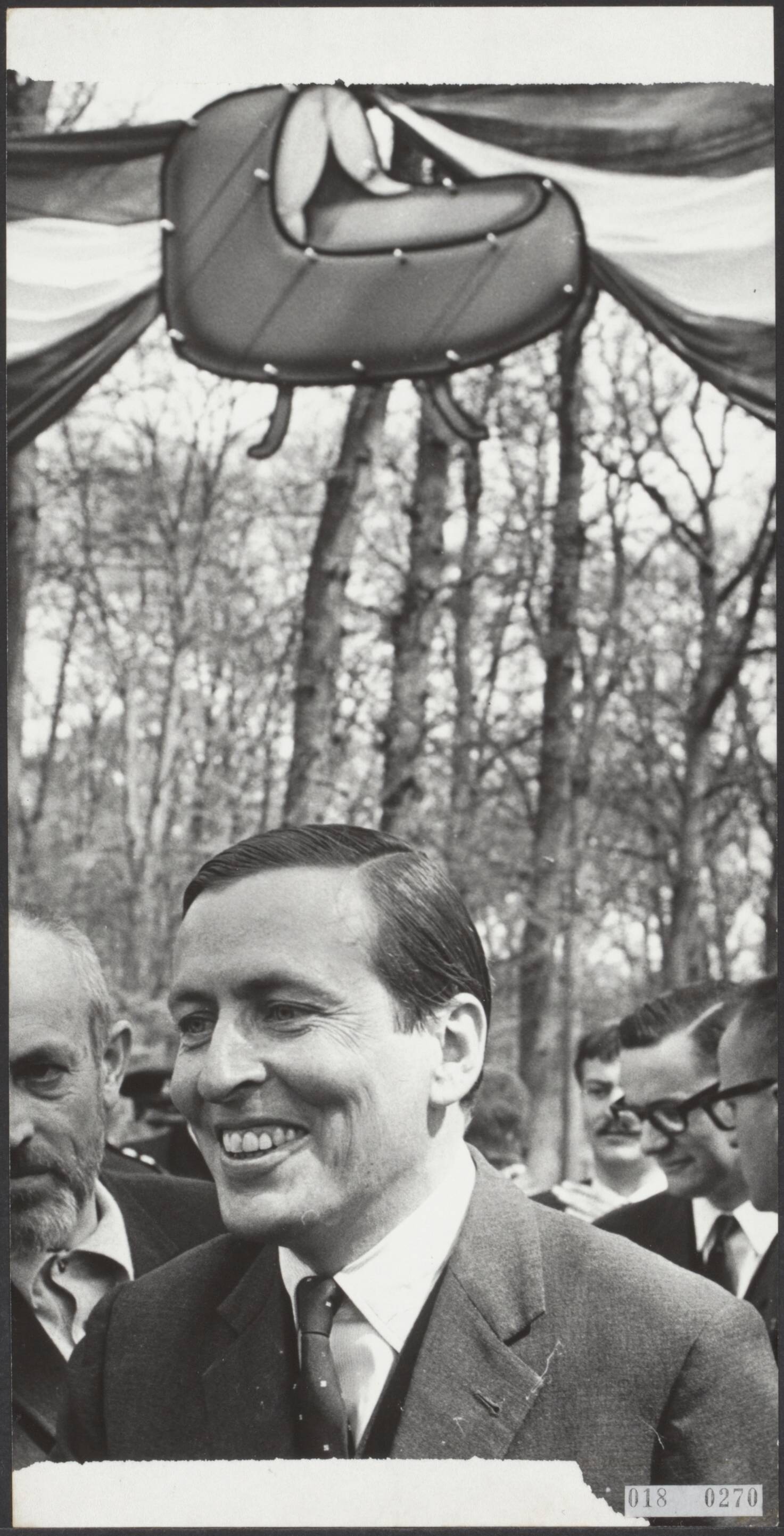Fotocollectie Elsevier Binnenland — Prins Claus kwam vanmiddag in Lage Vuursche even buiten de poort van kasteel Drakensteyn om voor de televisie een korte verklaring af te leggen. Boven de poort hing een passende versiering: een wiegje — Foto: Koch, Eric / Anefo. Licentie: Set B: Rechtenvrij / Publiek Domein. Bron: Nationaal Archief. http://hdl.handle.net/10648/adc1bf1e-d0b4-102d-bcf8-003048976d84