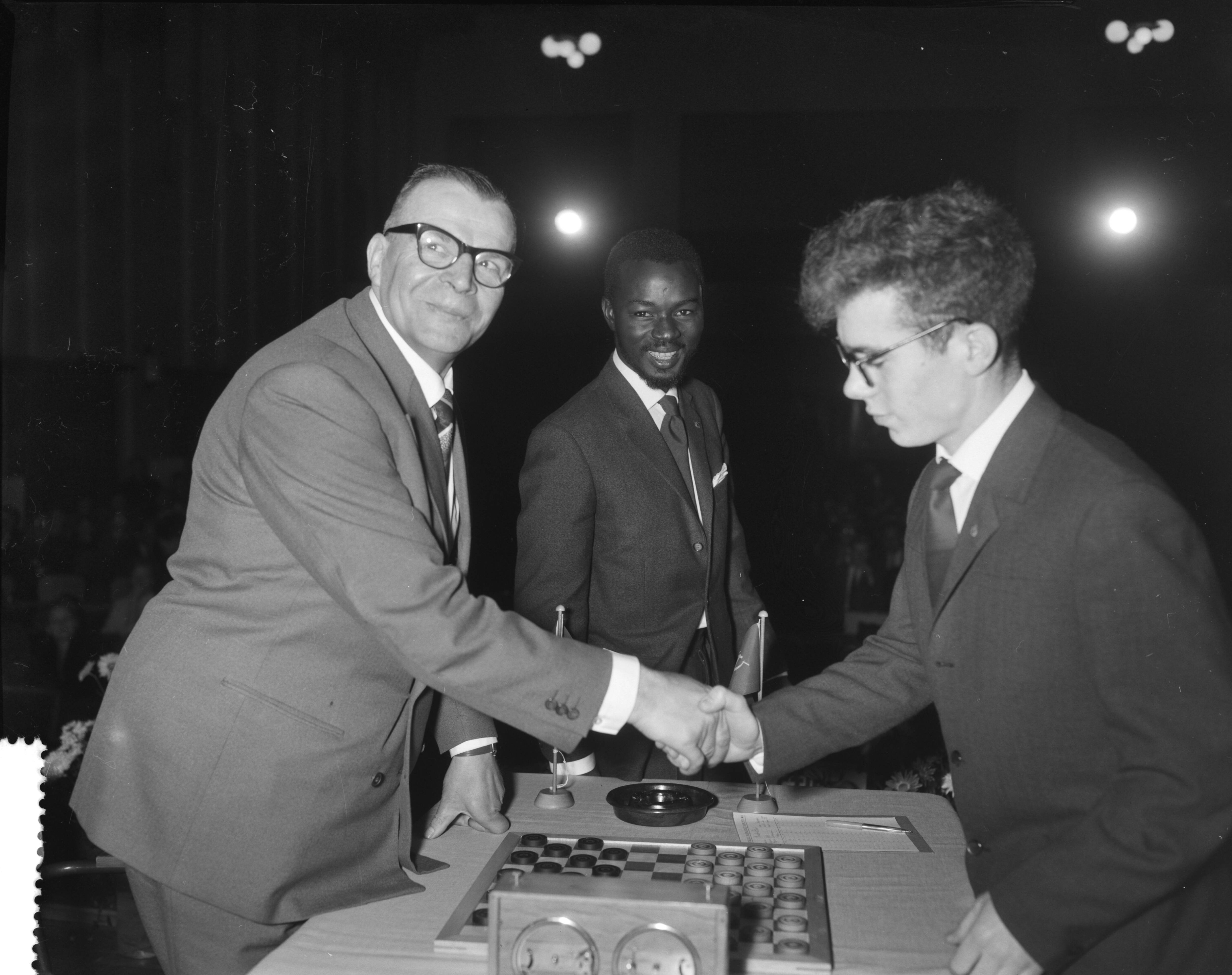 Fotocollectie Anefo — Laatste ronde wereldkampioenschap dammen in het Concertgebouw te Haarlem. Baris Dukel feliciteert wereldkampioen Tsjegolev (rechts). Baby Sy kijkt toe — Foto: Bilsen, Joop van / Anefo. Licentie: Set B: Rechtenvrij / Publiek Domein. Bron: Nationaal Archief. http://hdl.handle.net/10648/a9bf3f22-d0b4-102d-bcf8-003048976d84