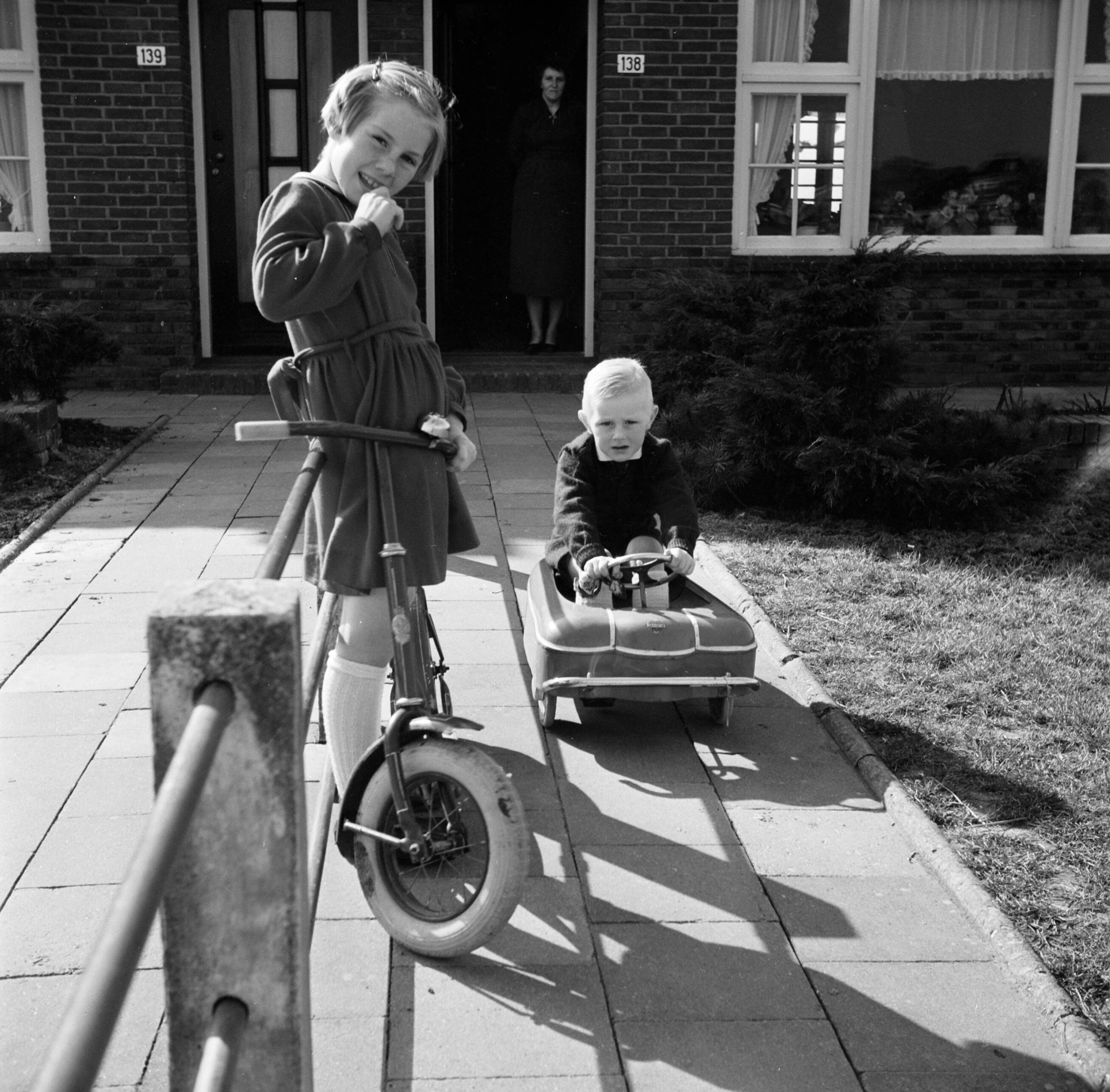 Fotocollectie Van de Poll — Marietje, de achtjarige dochter van Gerard, op haar autoped in de voortuin, samen met haar vierjarige broertje Gerrie. Haar moeder kijkt toe in de deuropening — Foto: Poll, Willem van de. Licentie: Set B: Rechtenvrij / Publiek Domein. Bron: Nationaal Archief. http://hdl.handle.net/10648/aecccdc2-d0b4-102d-bcf8-003048976d84