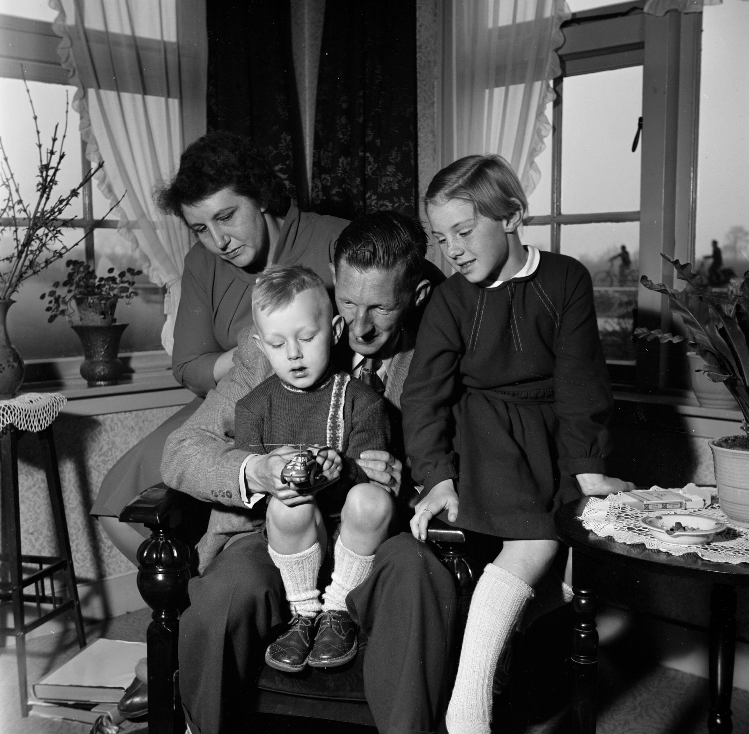 Fotocollectie Van de Poll — Gerard, zijn vrouw en hun beide kinderen in de huiskamer terwijl ze naar een stuk speelgoed kijken — Foto: Poll, Willem van de. Licentie: Set B: Rechtenvrij / Publiek Domein. Bron: Nationaal Archief. http://hdl.handle.net/10648/aeccd5e2-d0b4-102d-bcf8-003048976d84