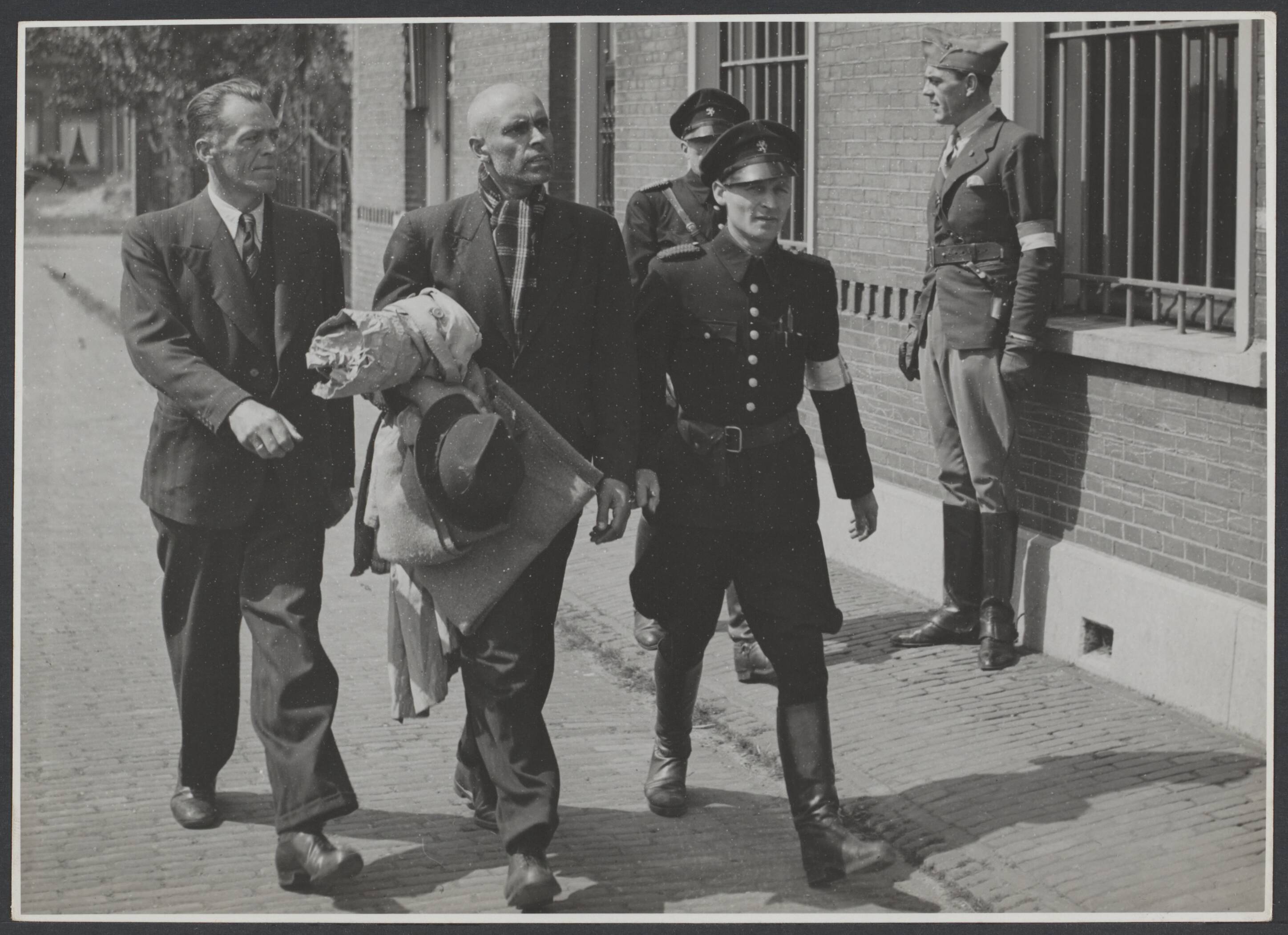 Collaboratie Nederland. — Haarlem [gearresteerde collaborateur wordt afgevoerd naar een met tralies voorzien gebouw. Tegen de muur een man in een soort fantasieuniform] — Foto: Nationaal Foto Persbureau (Stevens & Magielsen) / Rechtenvrij. Licentie: Set B: Rechtenvrij / Publiek Domein. Bron: Nationaal Archief. http://hdl.handle.net/10648/de0a3517-3dc8-c8e6-57ab-4544fb99128b