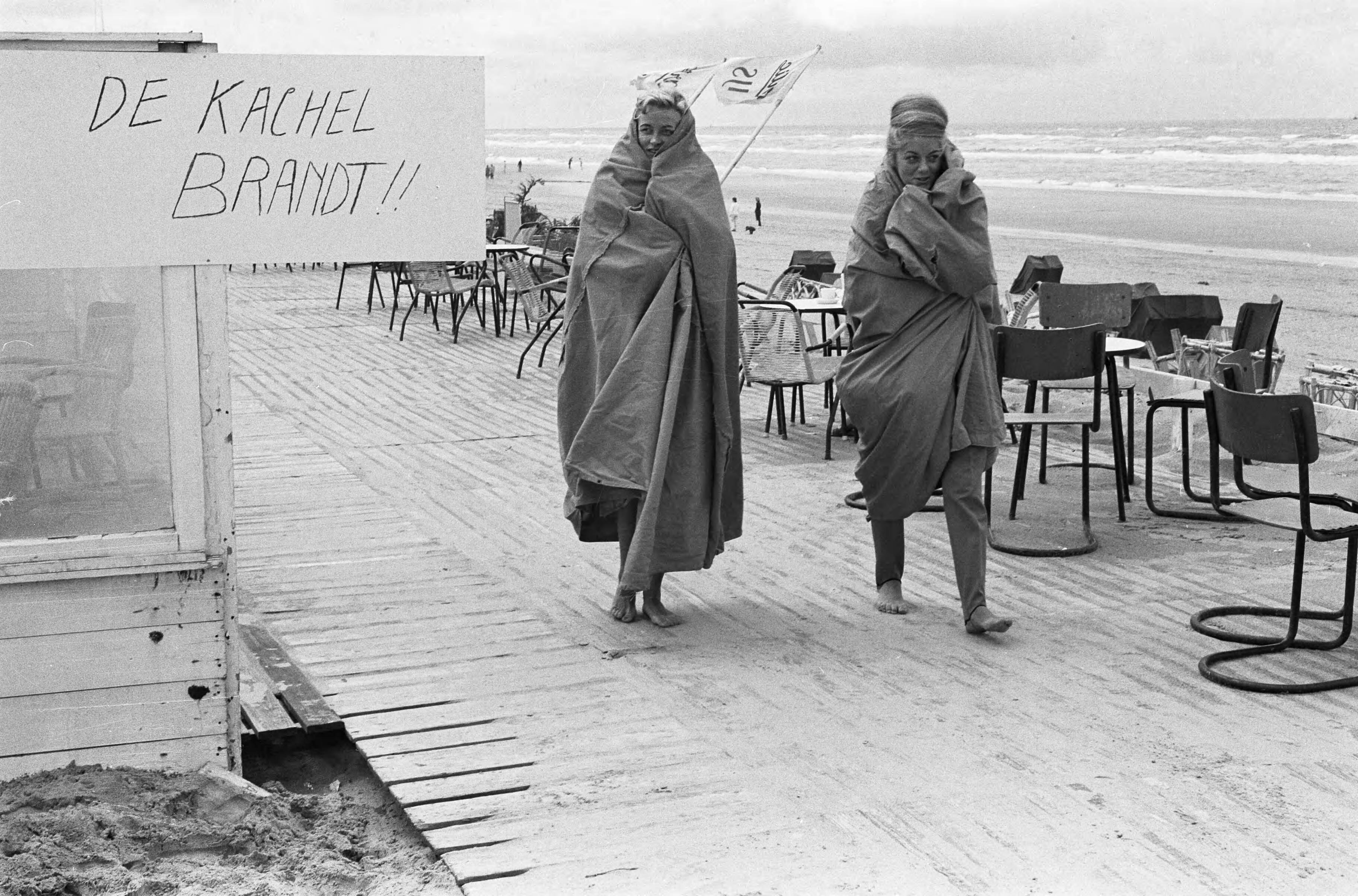 Fotocollectie Anefo — Slecht weer strand Zandvoort. Trieste strandterrasjes met bord met opschrift "De kachel brandt!!" en twee vrouwelijke badgasten gehuld in tentdoeken — Foto: Pot, Harry / Anefo. Licentie: Set B: Rechtenvrij / Publiek Domein. Bron: Nationaal Archief. http://hdl.handle.net/10648/aa0f5d22-d0b4-102d-bcf8-003048976d84