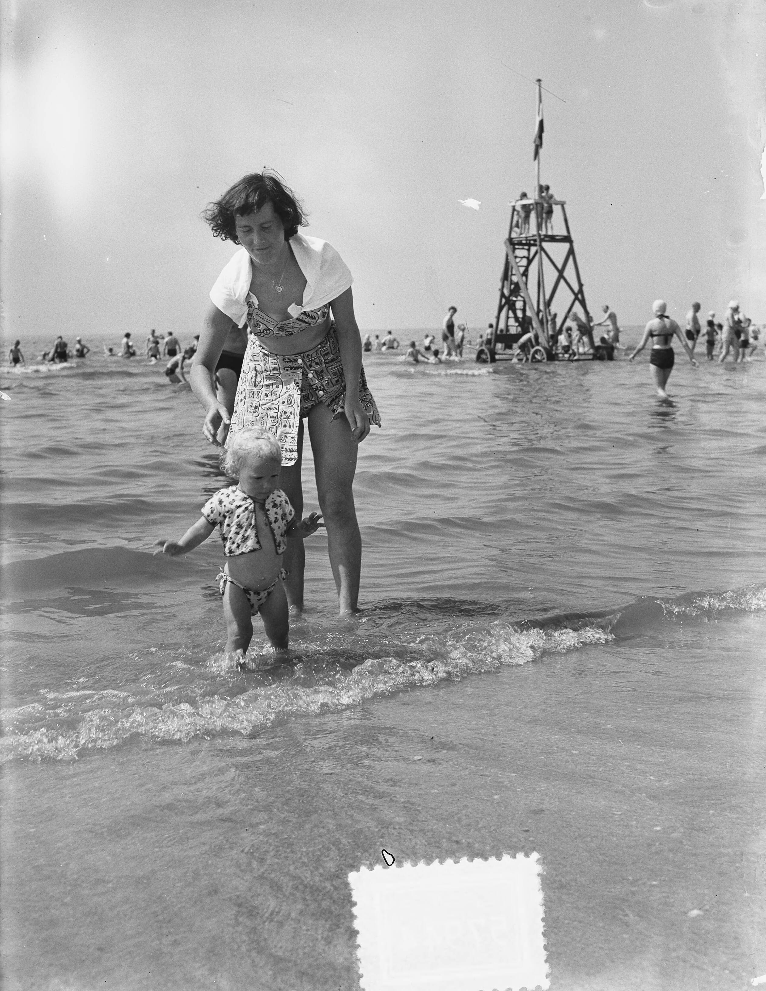 Fotocollectie Anefo — Zomerplaatjes. Drukte op het strand van Zandvoort. Vrouw met kind in het water — Foto: Rossem, Wim van / Anefo. Licentie: Set B: Rechtenvrij / Publiek Domein. Bron: Nationaal Archief. http://hdl.handle.net/10648/a90e3466-d0b4-102d-bcf8-003048976d84