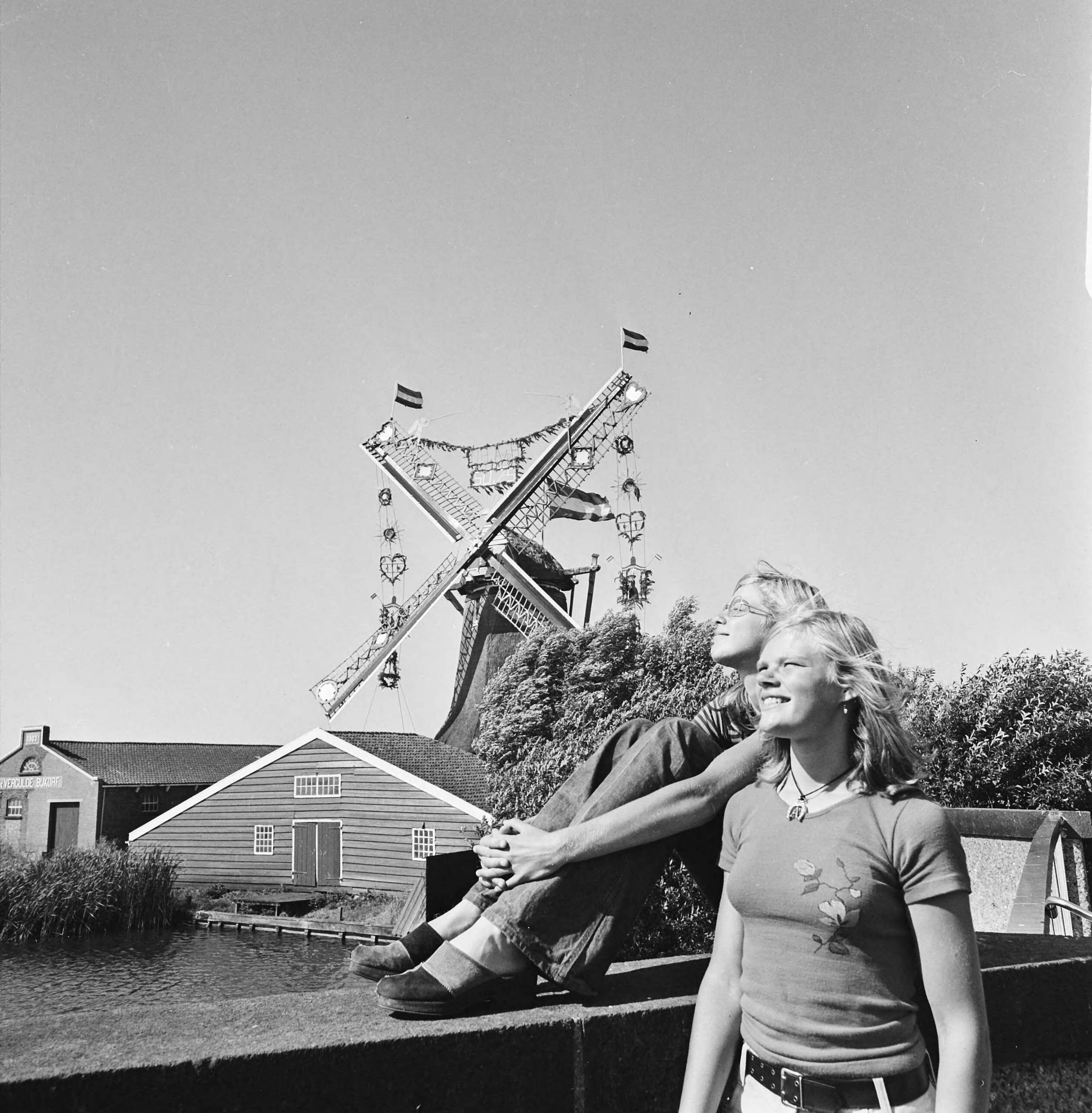 Molen in bruidstooi in Westzaan i.v.m. een tentoonstelling — Twee jongeren met op de achtergrond de met vlaggen en slingers versierde molen — Foto: Mieremet, Rob / Anefo. Licentie: Set B: Rechtenvrij / Publiek Domein. Bron: Nationaal Archief. http://hdl.handle.net/10648/ac6ee93e-d0b4-102d-bcf8-003048976d84