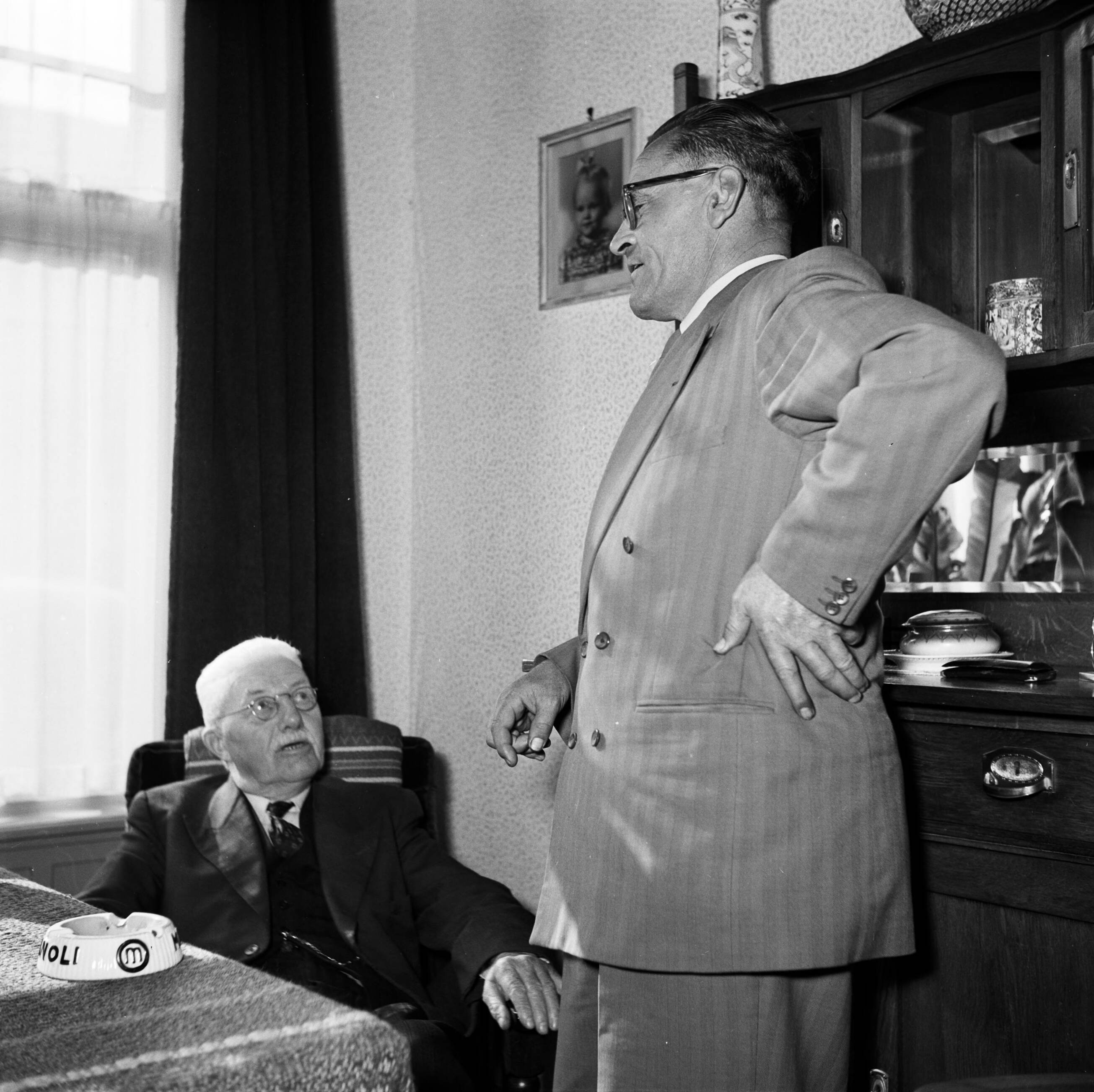 Fotocollectie Van de Poll — Gerard de Jongs vader, gezeten in een gemakkelijke stoel, in gesprek met Ynse Postma. Op tafel een grote asbak — Foto: Poll, Willem van de. Licentie: Set B: Rechtenvrij / Publiek Domein. Bron: Nationaal Archief. http://hdl.handle.net/10648/aecce7f8-d0b4-102d-bcf8-003048976d84