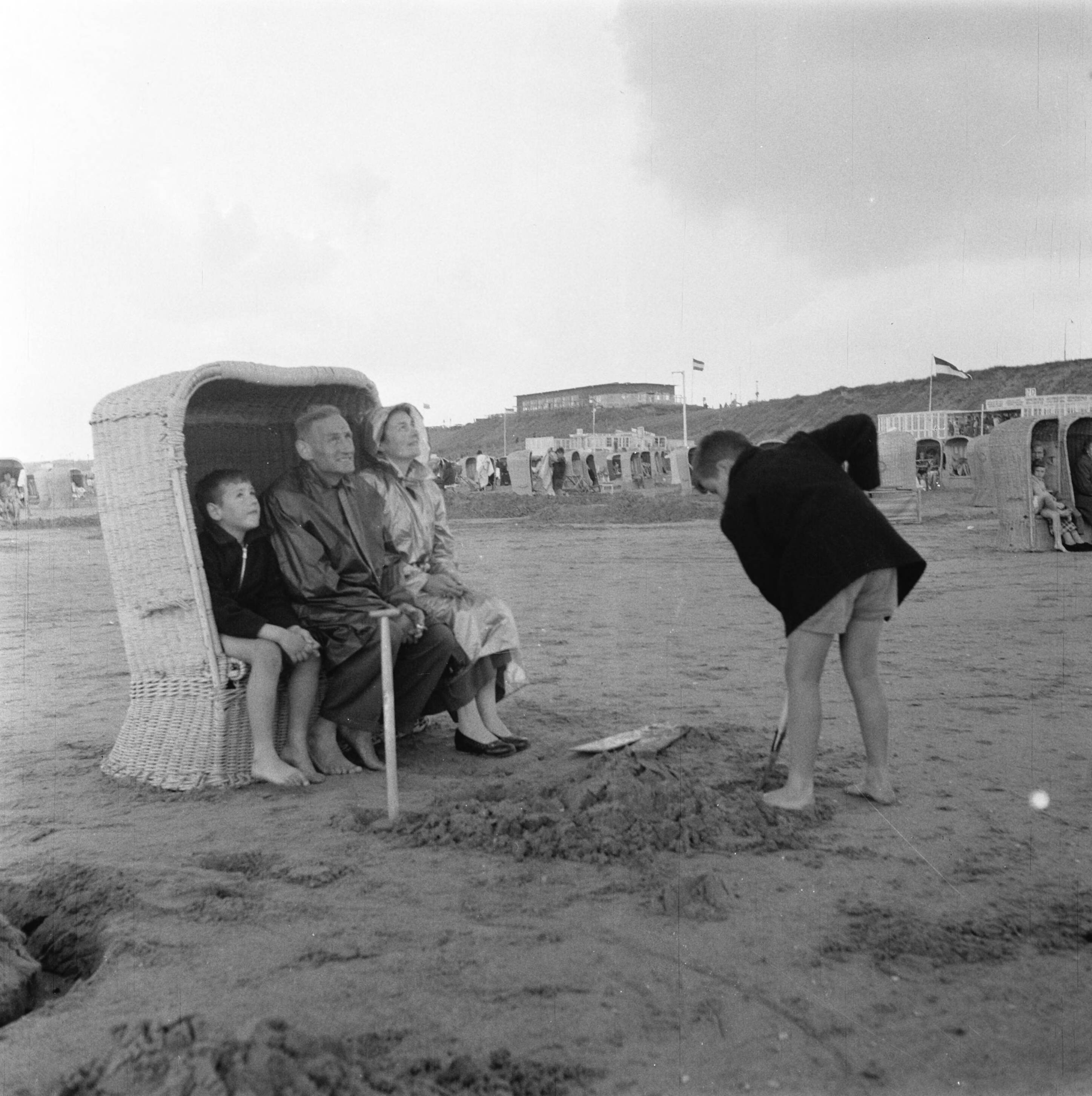 Fotocollectie Anefo — Plaatjes Zandvoort [gezin zit in strandstoel met slecht weer. Een zoon schept zand] — Foto: Noske, J.D. / Anefo. Licentie: Set B: Rechtenvrij / Publiek Domein. Bron: Nationaal Archief. http://hdl.handle.net/10648/a94dadee-d0b4-102d-bcf8-003048976d84
