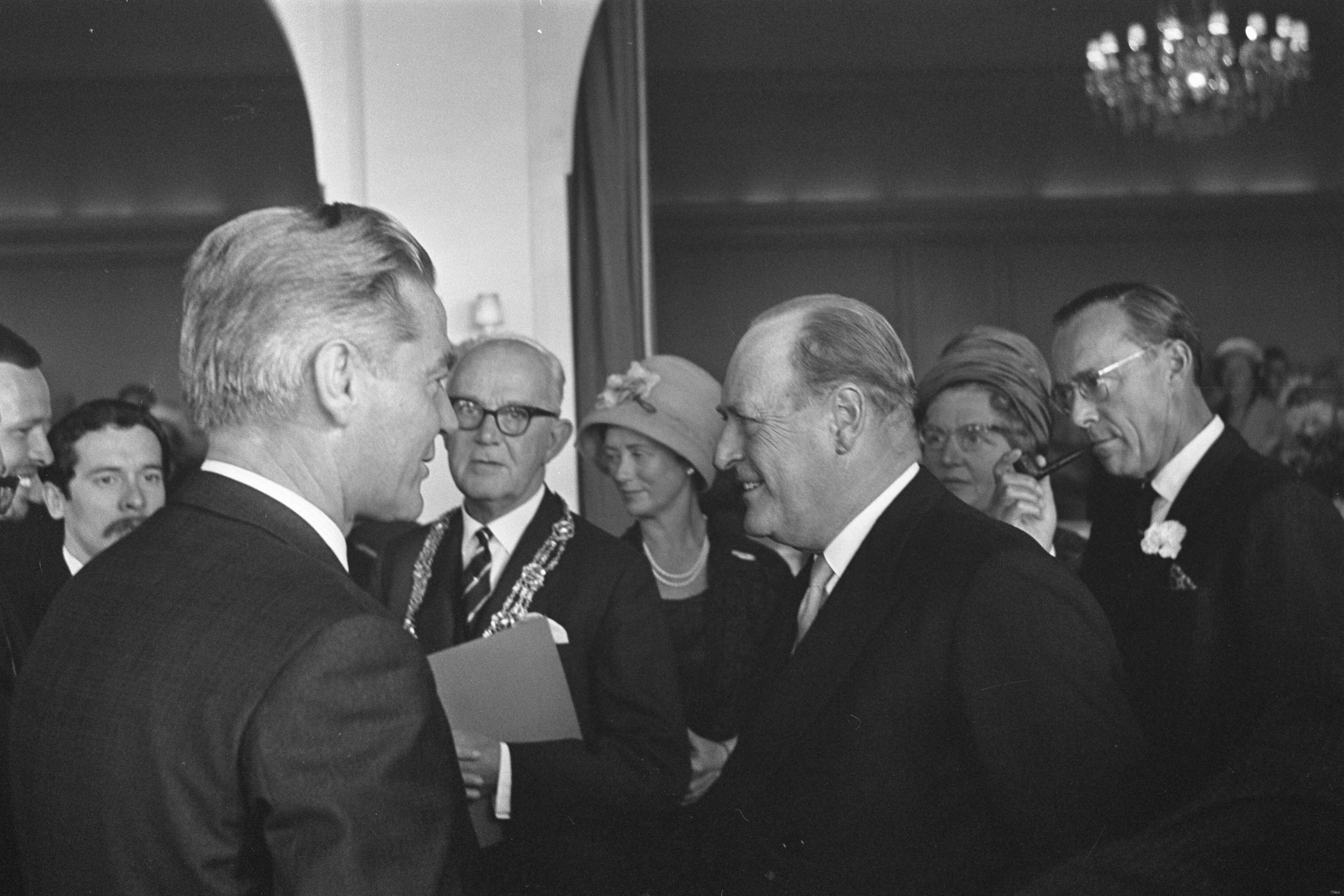 Staatsbezoek van koning Olav van Noorwegen aan Nederland — Bezoek koning Olav van Noorwegen aan Den Haag. Koning Olav in gesprek met filmer Bert Haanstra en dhr. Kolfschoten, burgemeester van Den Haag — Foto: Nijs, Jac. de / Anefo. Licentie: Set B: Rechtenvrij / Publiek Domein. Bron: Nationaal Archief. http://hdl.handle.net/10648/aa7873ac-d0b4-102d-bcf8-003048976d84