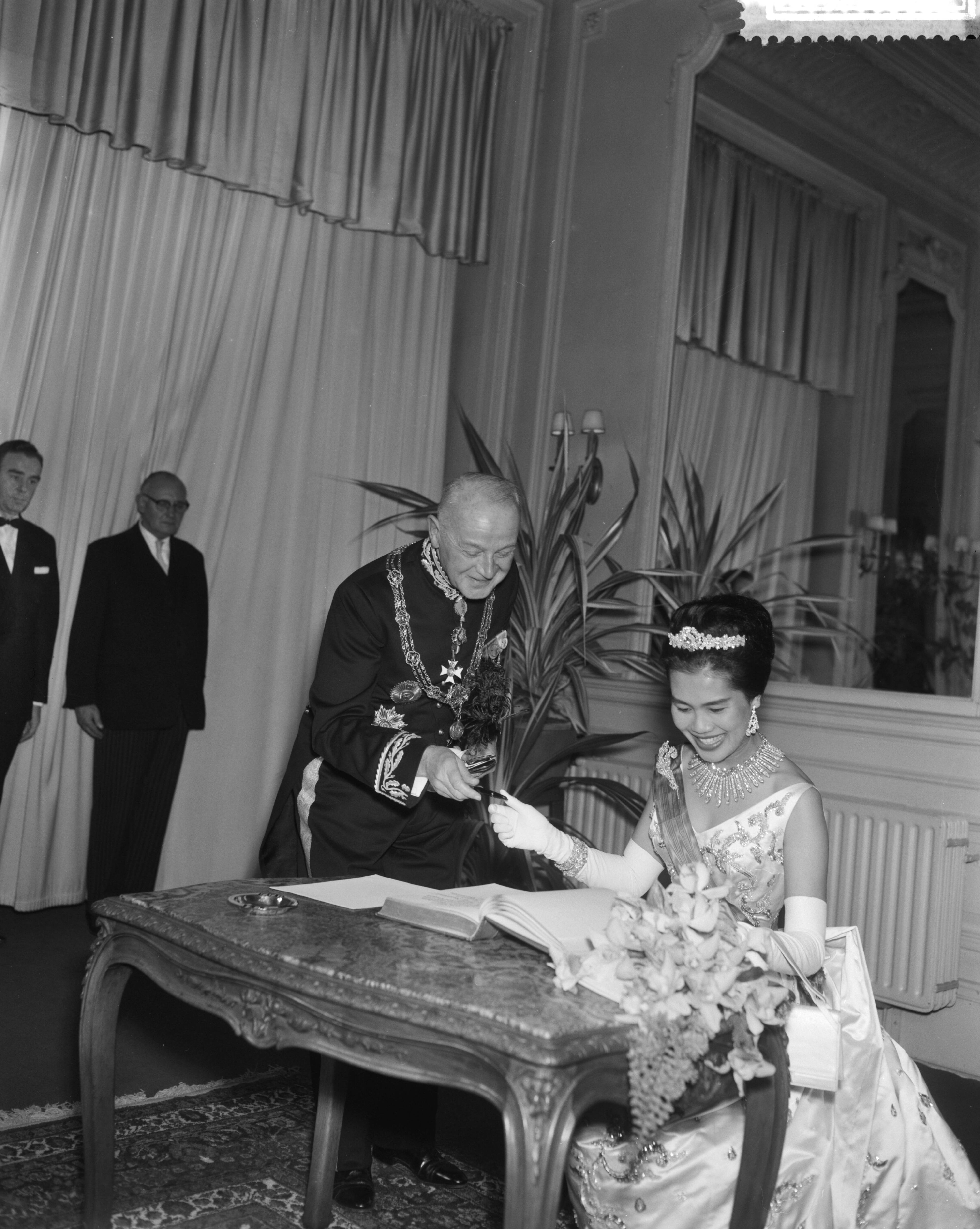 Fotocollectie Anefo — Galaconcert door Koninklijke Militaire Kapel in de Kurzaal te Den Haag , Koningin Sirikit tekent gastenboek, links burgemeester Kolfschoten — Foto: Bilsen, Joop van / Anefo. Licentie: Set B: Rechtenvrij / Publiek Domein. Bron: Nationaal Archief. http://hdl.handle.net/10648/a9bca3fc-d0b4-102d-bcf8-003048976d84