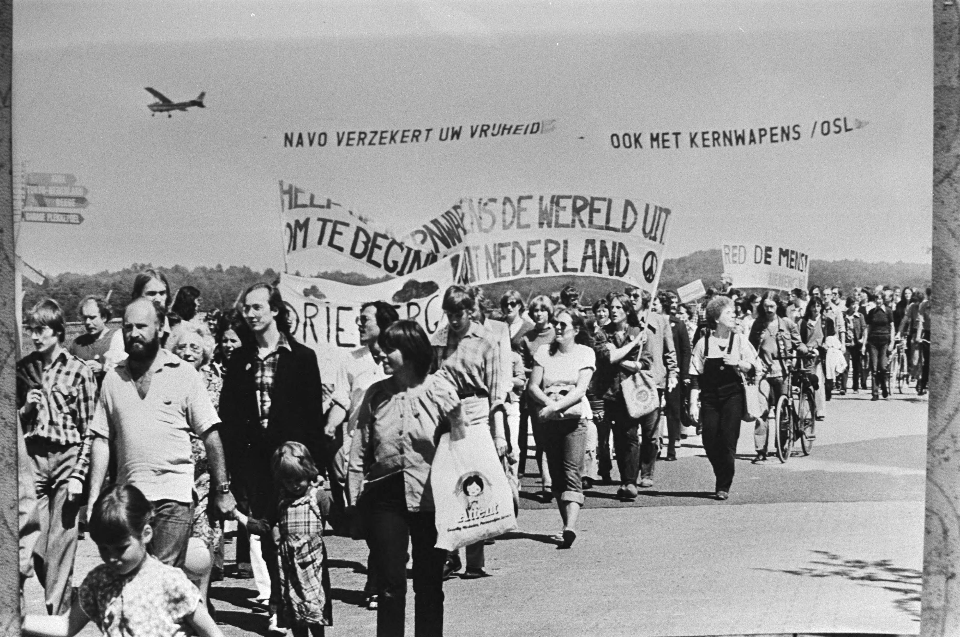 Demonstratie in Soesterberg tegen kernbewapening en opslag van kernwapens op vliegveld Soesterberg — Op de spandoeken staat o.a. "Alle kernwapens de wereld uit - om te beginnen uit Nederland". Tegendemonstratie in de lucht: op de sleep van een reclamevliegtuigje staat te lezen "NAVO verzekert uw vrijheid - ook met kernwapens" — Foto: Croes, Rob C. / Anefo. Licentie: Set B: Rechtenvrij / Publiek Domein. Bron: Nationaal Archief. http://hdl.handle.net/10648/acca75b0-d0b4-102d-bcf8-003048976d84
