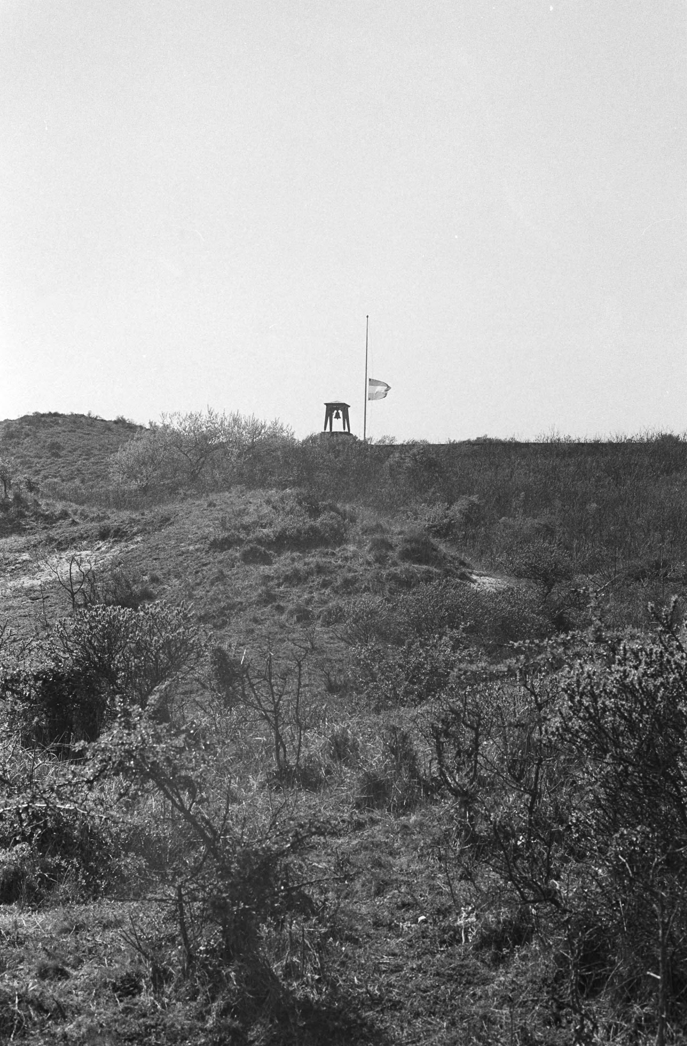 Jaarlijkse herdenking van 1940-1945 op de erebegraafplaats te Overveen — Zicht vanuit de duinen op de toren met klok en de vlag halfstok — Foto: Fotograaf Onbekend / Anefo. Licentie: Set B: Rechtenvrij / Publiek Domein. Bron: Nationaal Archief. http://hdl.handle.net/10648/abbe9b88-d0b4-102d-bcf8-003048976d84