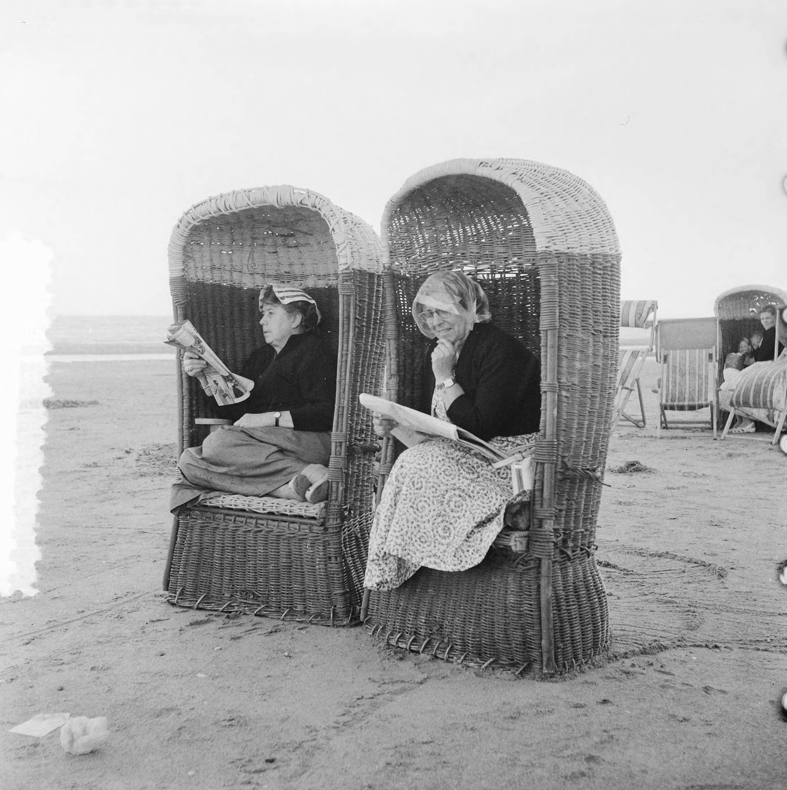 Fotocollectie Anefo — Plaatjes Zandvoort [twee vrouwen lezen de krant in rieten strandstoelen tijdens slecht weer] — Foto: Noske, J.D. / Anefo. Licentie: Set B: Rechtenvrij / Publiek Domein. Bron: Nationaal Archief. http://hdl.handle.net/10648/ad77d5d4-d0b4-102d-bcf8-003048976d84