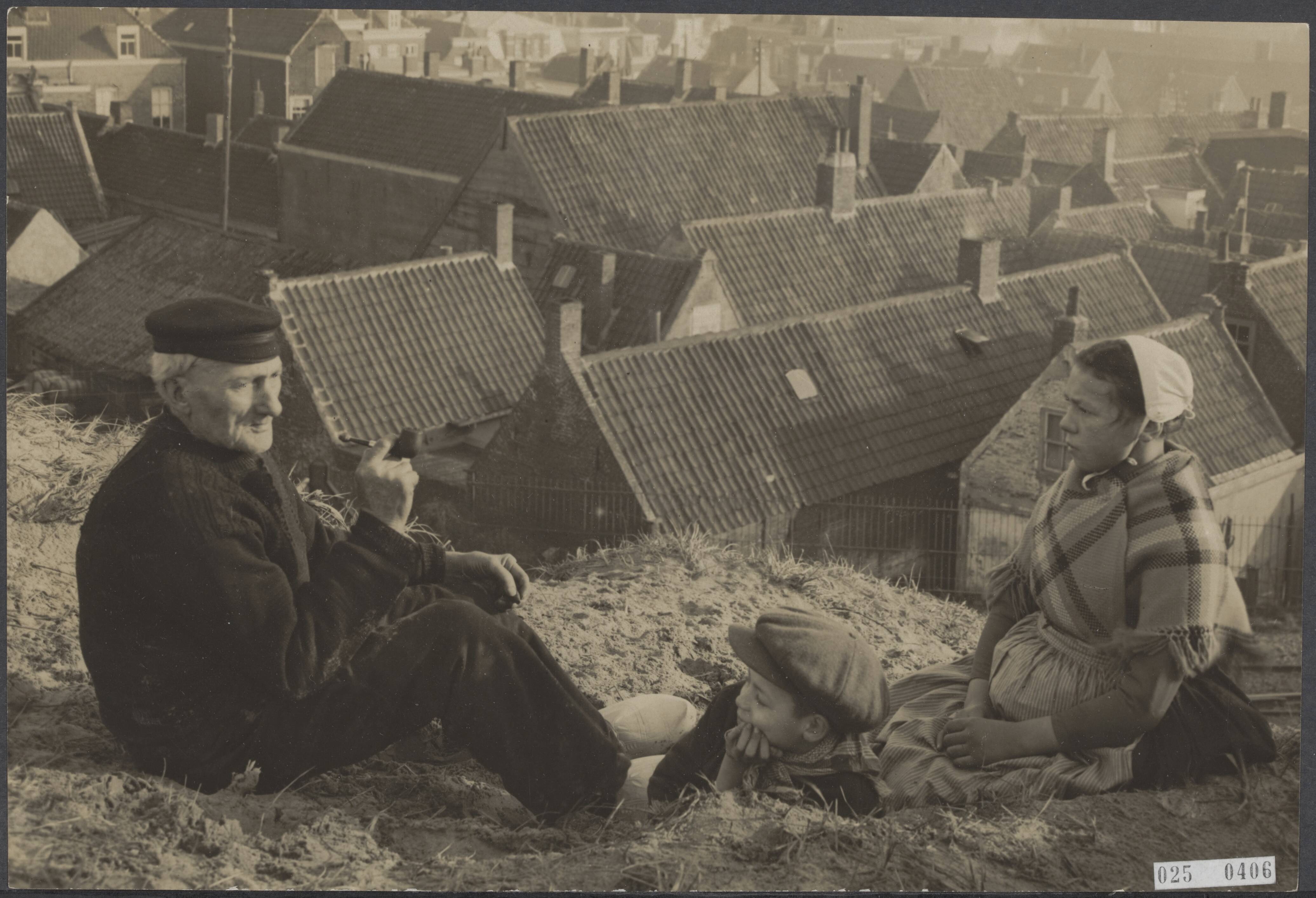 Fotocollectie Elsevier Binnenland — Oudere man, jongen en vrouw zitten op een duin met zicht op een dorp [locatie waarschijnlijk Scheveningen] — Foto: Onbekend / Publiek Domein of Rechtenvrij. Licentie: Set B: Rechtenvrij / Publiek Domein. Bron: Nationaal Archief. http://hdl.handle.net/10648/adda728e-d0b4-102d-bcf8-003048976d84
