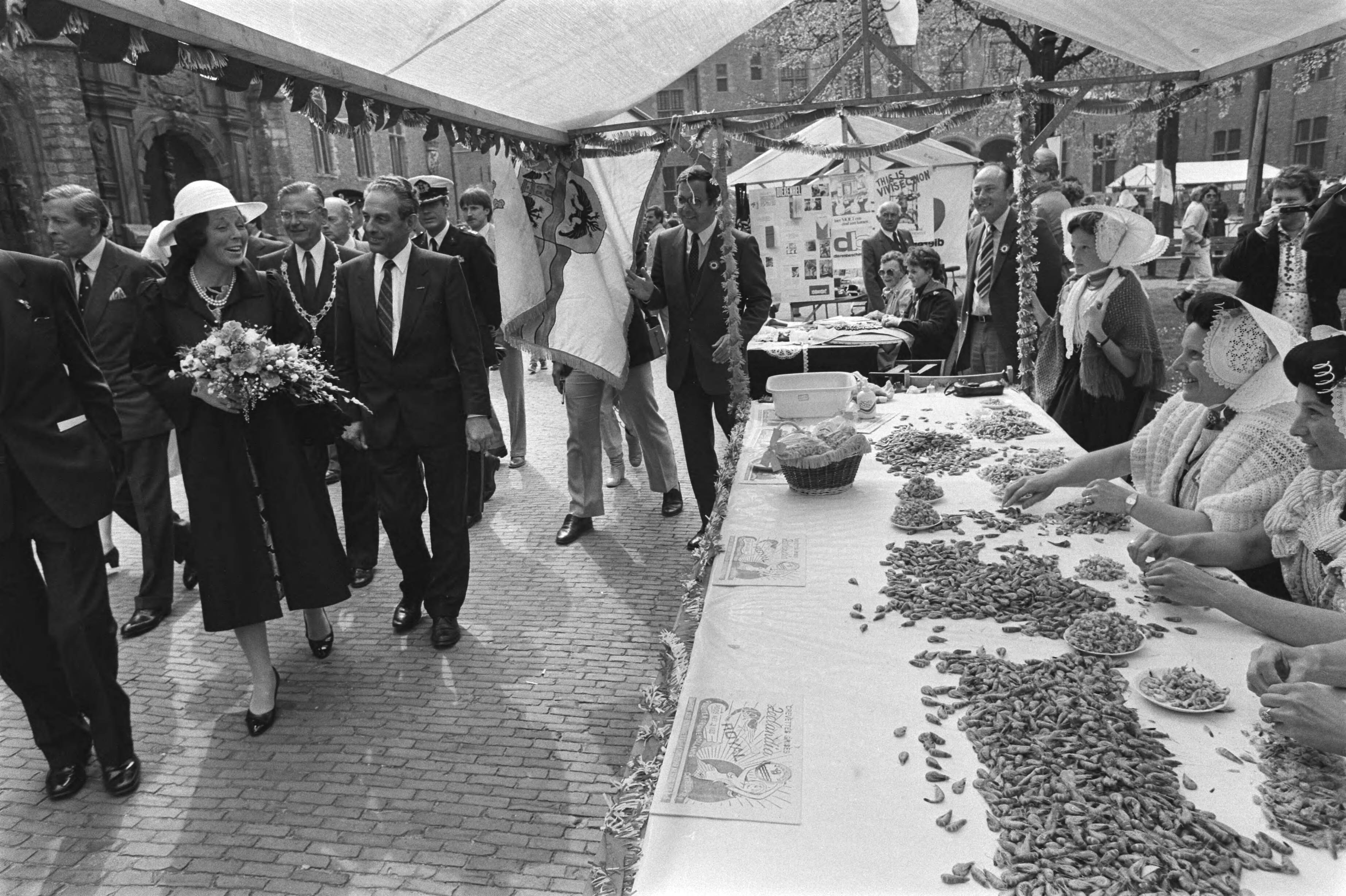 Fotocollectie Anefo — Kon. Beatrix en Prins Claus bij Nat. herdenkingsmanifestatie in Middelburg; Koningin Beatrix en Prins Claus lopen langs kraam met garnalenpelsters / — Foto: Bogaerts, Rob / Anefo. Licentie: Set B: Rechtenvrij / Publiek Domein. Bron: Nationaal Archief. http://hdl.handle.net/10648/ad385864-d0b4-102d-bcf8-003048976d84