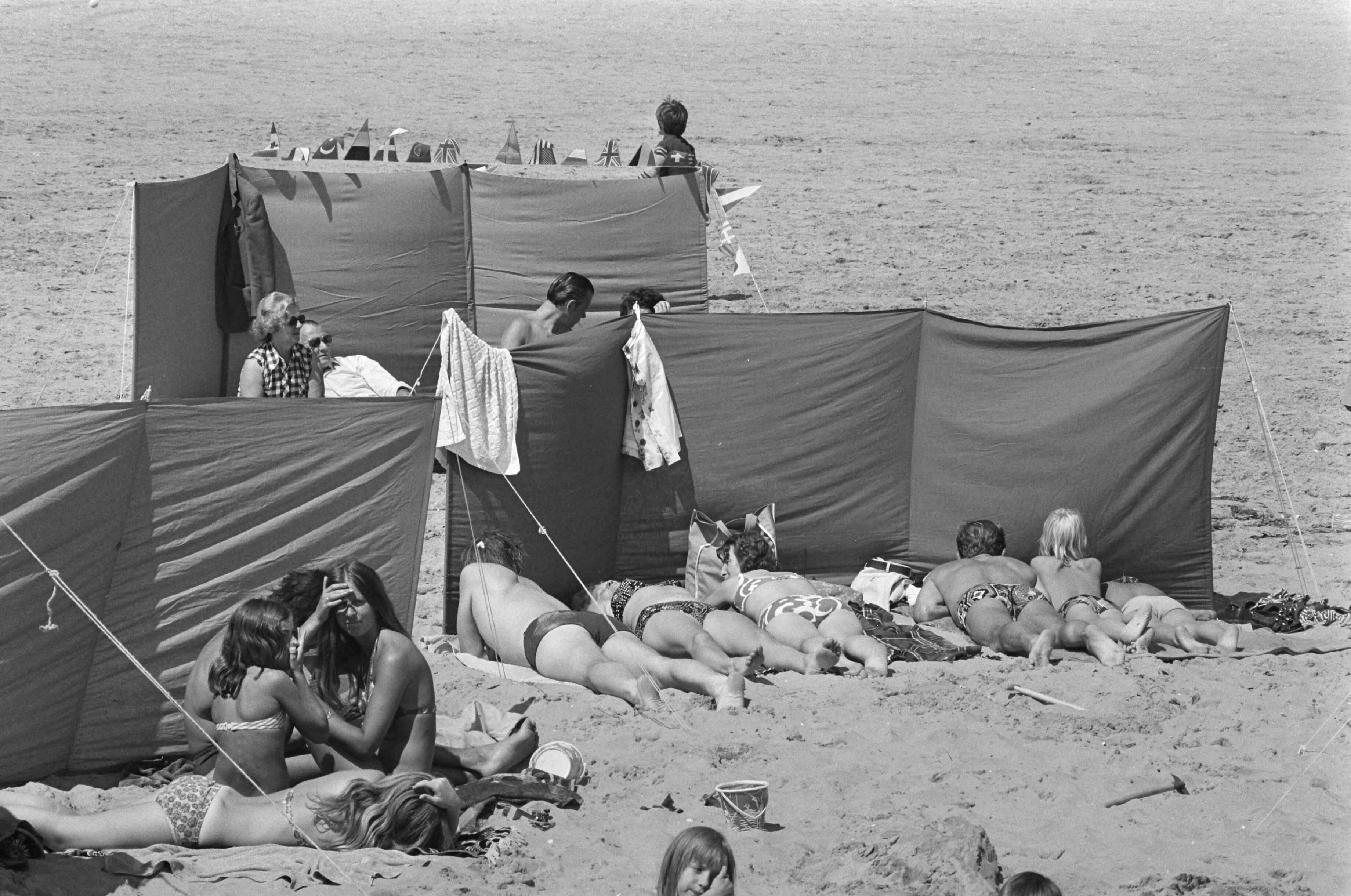 Fotocollectie Anefo — Drukte op Scheveningse strand; rijtje zonnebaders achter windscherm — Foto: Verhoeff, Bert / Anefo. Licentie: Set B: Rechtenvrij / Publiek Domein. Bron: Nationaal Archief. http://hdl.handle.net/10648/ac50deee-d0b4-102d-bcf8-003048976d84