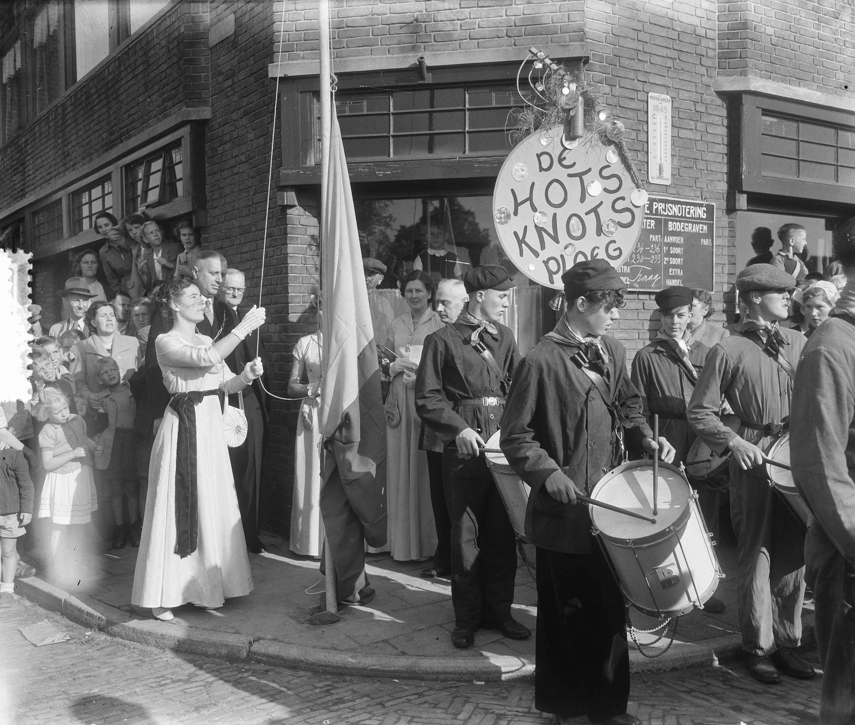 Fotocollectie Anefo — Kaaskoningin Lien I hijst een vlag tijdens haar rondgang door Bodegraven. Achter haar burgemeester J.J. Croles — Foto: Pot, Harry / Anefo. Licentie: Set B: Rechtenvrij / Publiek Domein. Bron: Nationaal Archief. http://hdl.handle.net/10648/a8ff6120-d0b4-102d-bcf8-003048976d84