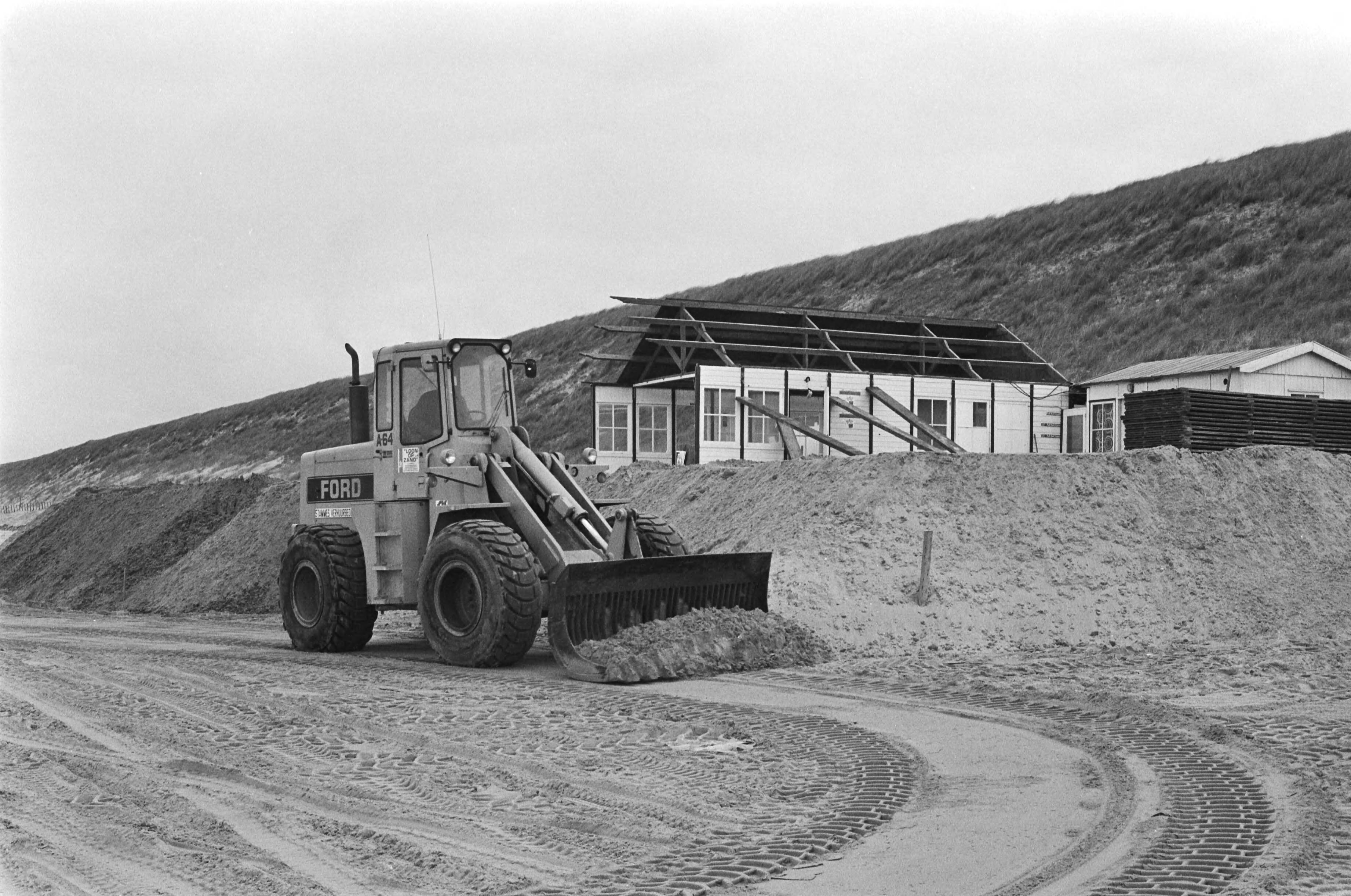 Fotocollectie Anefo — Herstelwerkzaamheden aan door stormschade getroffen strandtenten en paviljoens in Zandvoort; buldozer verhoogd zand bij strandhuisje — Foto: Dijk, Hans van / Anefo. Licentie: Set B: Rechtenvrij / Publiek Domein. Bron: Nationaal Archief. http://hdl.handle.net/10648/acdeb020-d0b4-102d-bcf8-003048976d84
