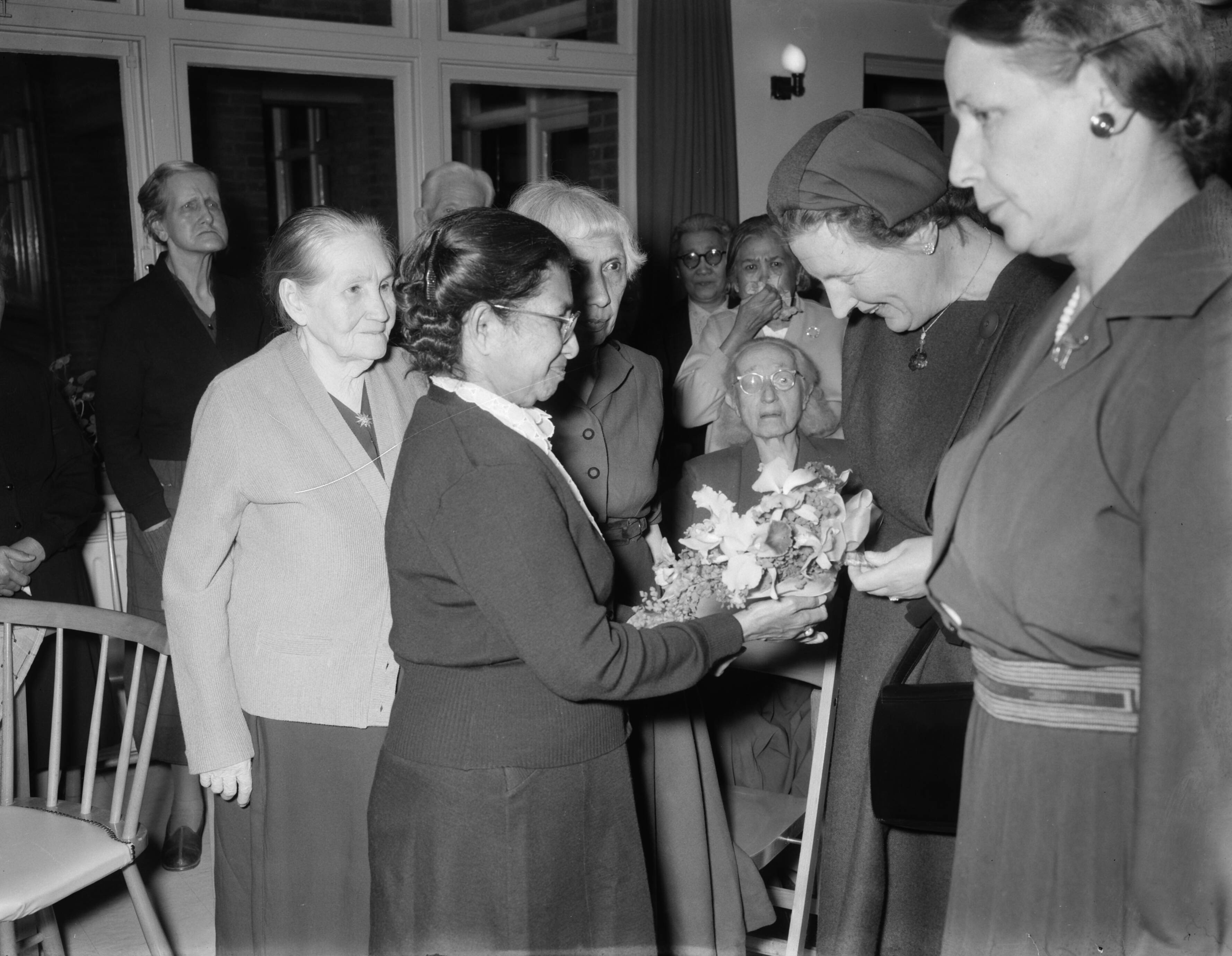 Fotocollectie Anefo — Koningin Juliana bezoekt bejaarde gerepatrieerden uit Indonesië in het protestantse tehuis voor bejaarden "Spathodea" te Zeist. De in 1909 geboren mevr. Julia Veronica Simons-Titaly biedt de koningin bij haar aankomst bloemen aan. Naast haar de 73e-jarige meverouw A.W.P. Geraths de Saussin, geboren in Palembang — Foto: Noske, J.D. / Anefo. Licentie: Set B: Rechtenvrij / Publiek Domein. Bron: Nationaal Archief. http://hdl.handle.net/10648/a973b6a6-d0b4-102d-bcf8-003048976d84