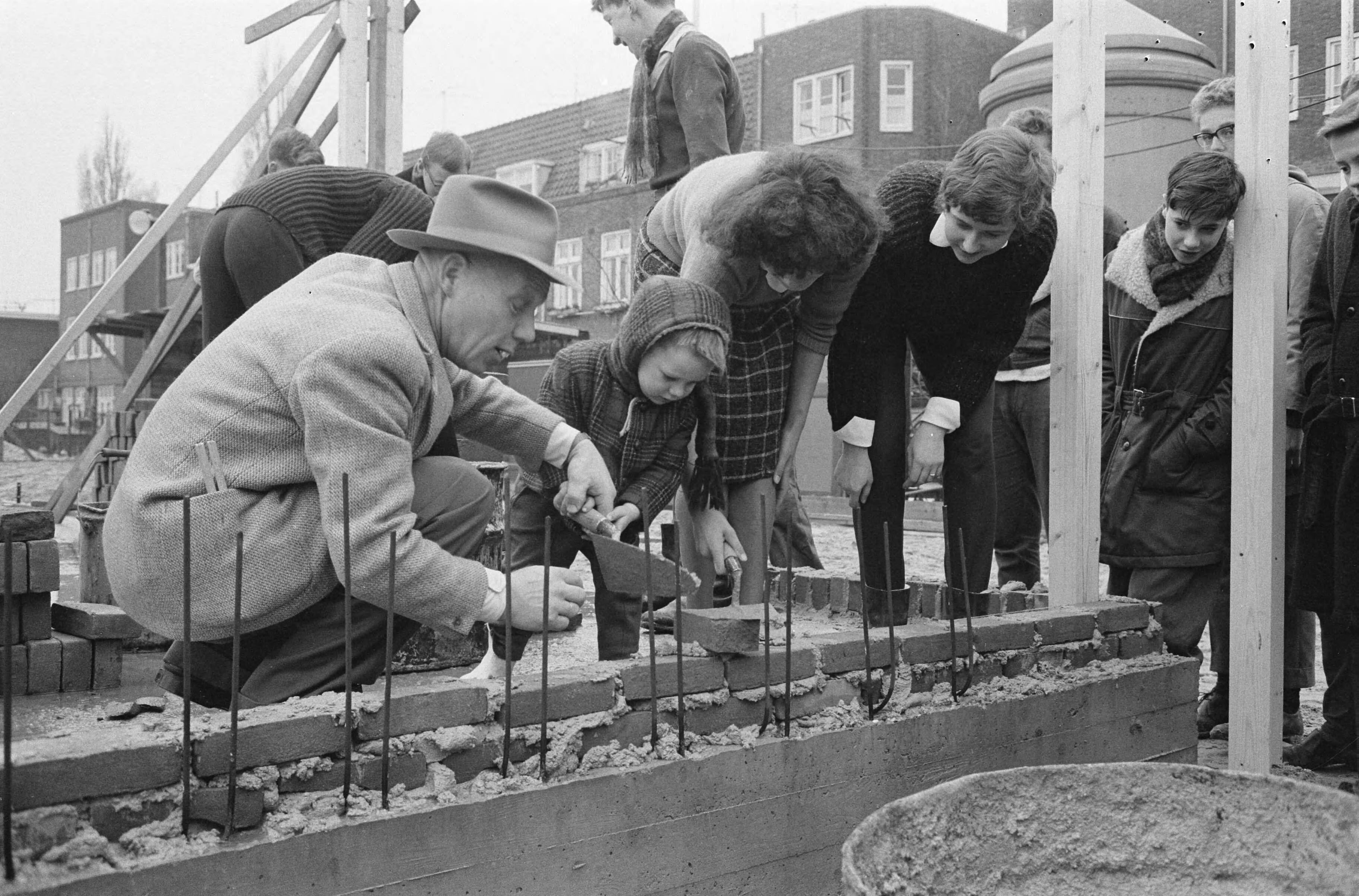 Fotocollectie Anefo — Leerlingen metselen eerste stenen van het Jeugdgebouw Noord aan de Meeuwenlaan te Amsterdam. Hansje Burger, 3 jaar, legt steentje onder leiding van heer Bakker — Foto: Broers, F.N. / Anefo. Licentie: Set B: Rechtenvrij / Publiek Domein. Bron: Nationaal Archief. http://hdl.handle.net/10648/aa236eac-d0b4-102d-bcf8-003048976d84