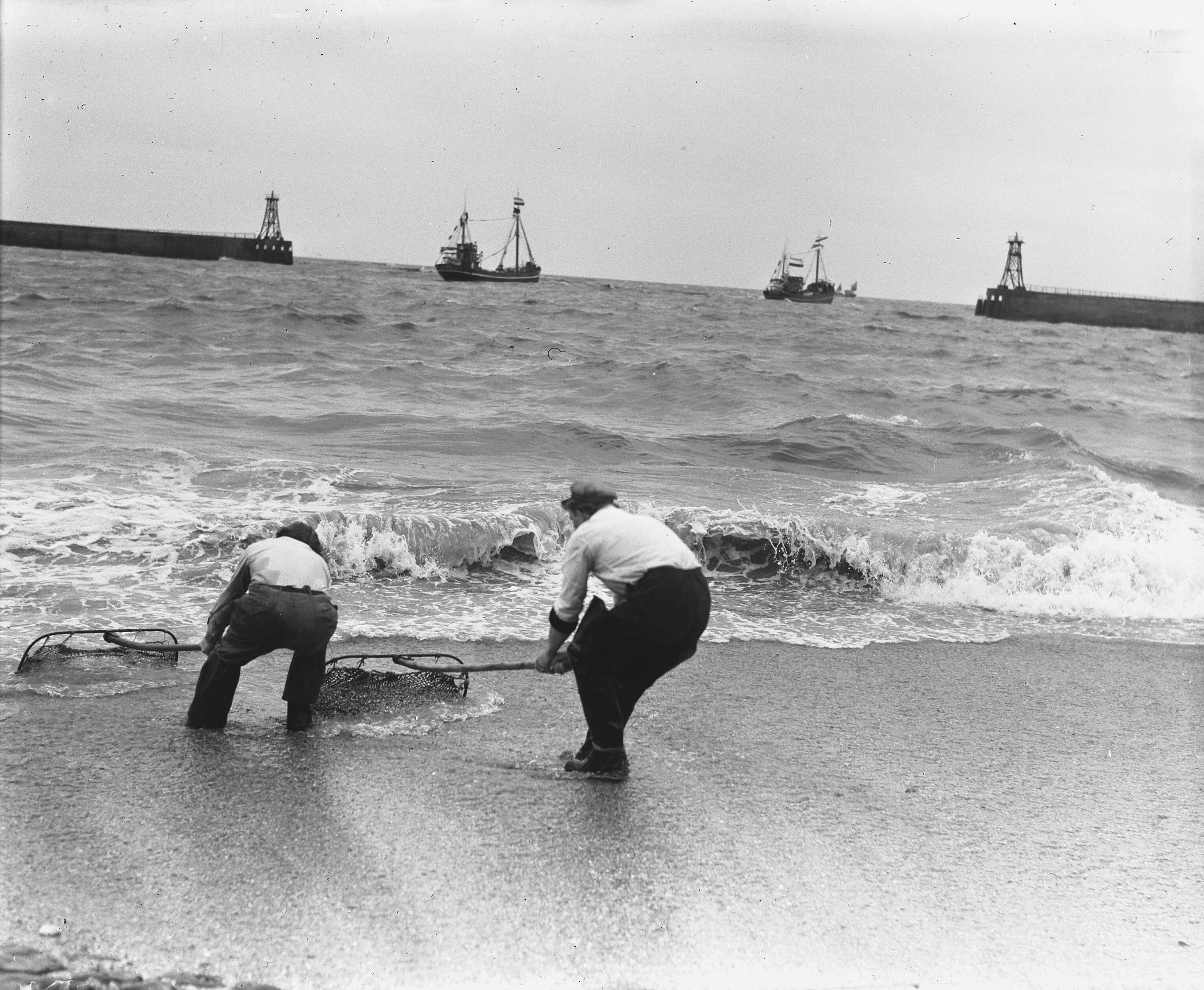 Uitvaren van de haringvloot in Scheveningen — Garnalenvissers op het strand. In de achtergrond de haringvloot — Foto: Koch, Eric / Anefo. Licentie: Set B: Rechtenvrij / Publiek Domein. Bron: Nationaal Archief. http://hdl.handle.net/10648/a97c4406-d0b4-102d-bcf8-003048976d84