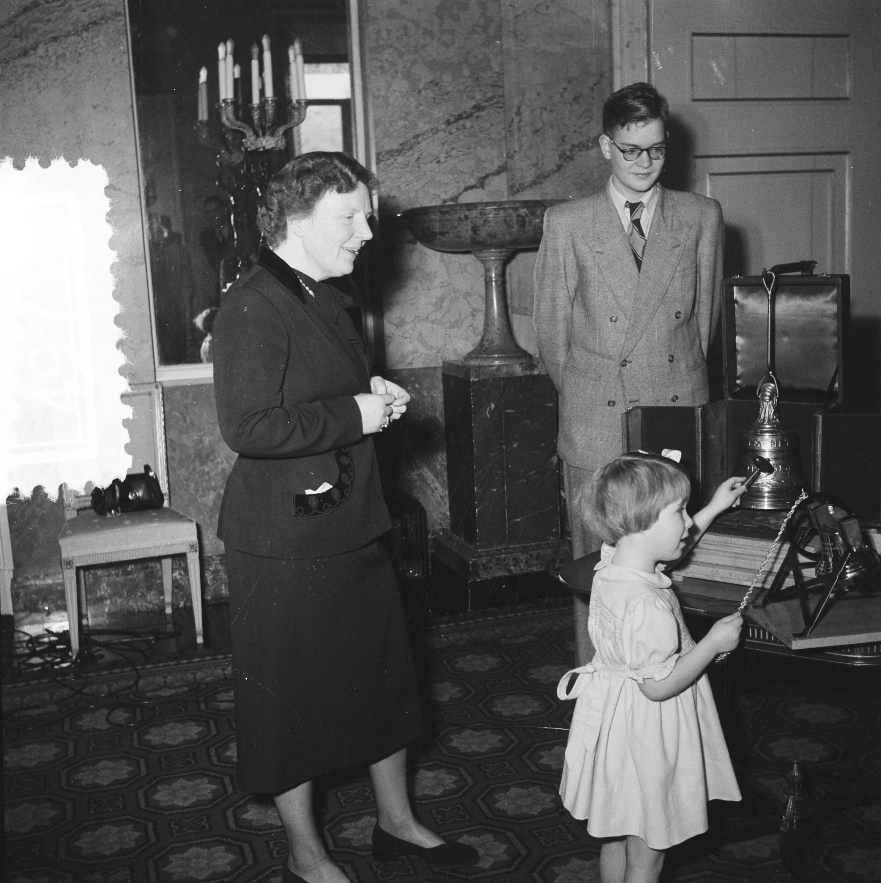 Aanbieding op paleis Soestdijk aan prinses Christina van de kleinste luidklok, het z.g. Marijke-klokje, behorend tot het carillon dat Nederland als dank aan de Verenigde Staten zal geven — Prinses Marijke (Christina) slaat tegen het klokje; links koningin Juliana, in het midden Rob Veenstra — Foto: Noske, J.D. / Anefo. Licentie: Set B: Rechtenvrij / Publiek Domein. Bron: Nationaal Archief. http://hdl.handle.net/10648/a8f599b0-d0b4-102d-bcf8-003048976d84
