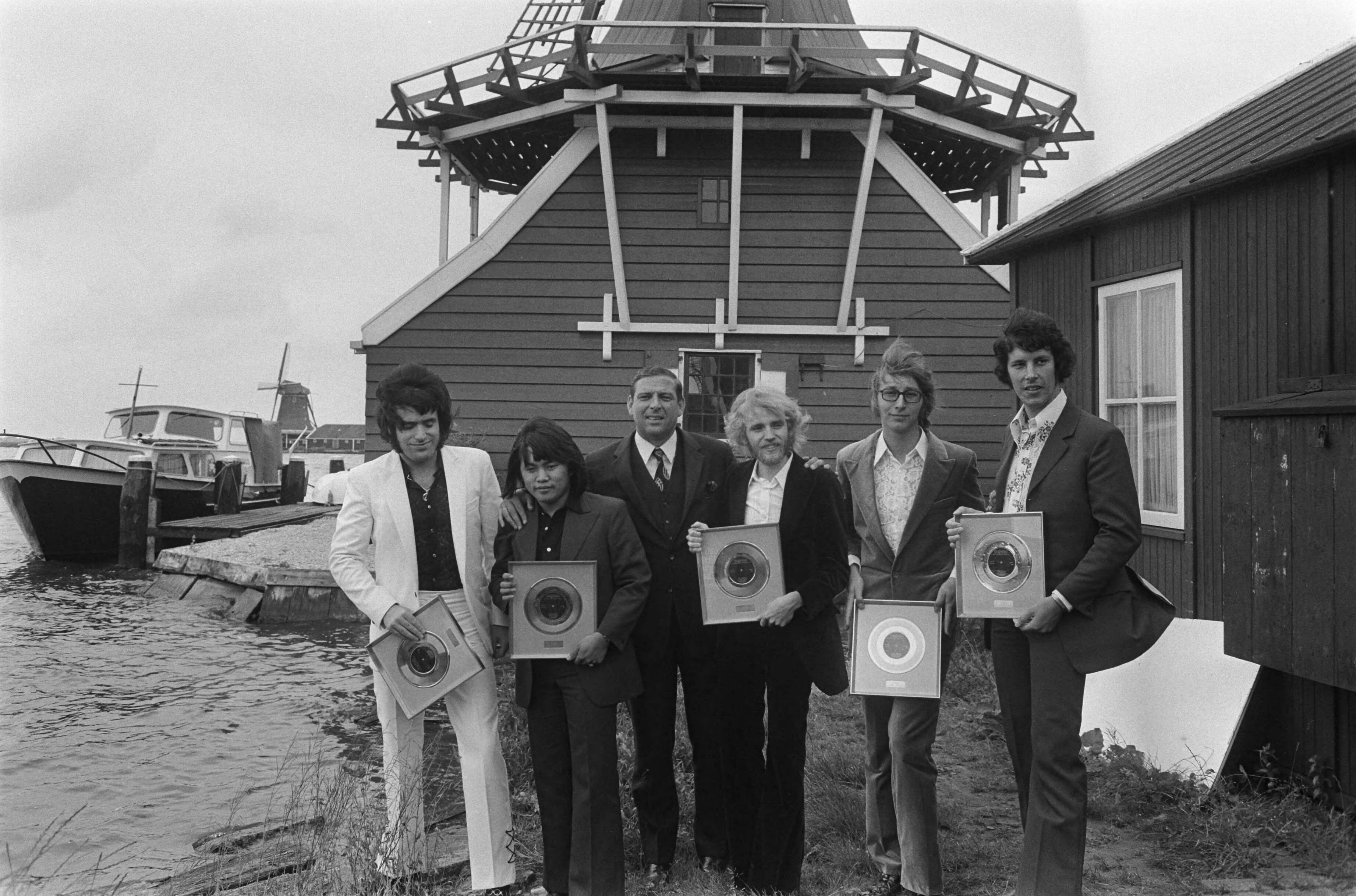 De Zaanse popgroep The George Baker Selection krijgt gouden plaat uitgereikt op de Zaanse Schans — Tegen het decor van de mosterdmolen De Huisman toot de band zijn trofee in gezelschap van een official van de platen maatschappij — Foto: Fotograaf Onbekend / Anefo. Licentie: Set B: Rechtenvrij / Publiek Domein. Bron: Nationaal Archief. http://hdl.handle.net/10648/ab9b40ca-d0b4-102d-bcf8-003048976d84