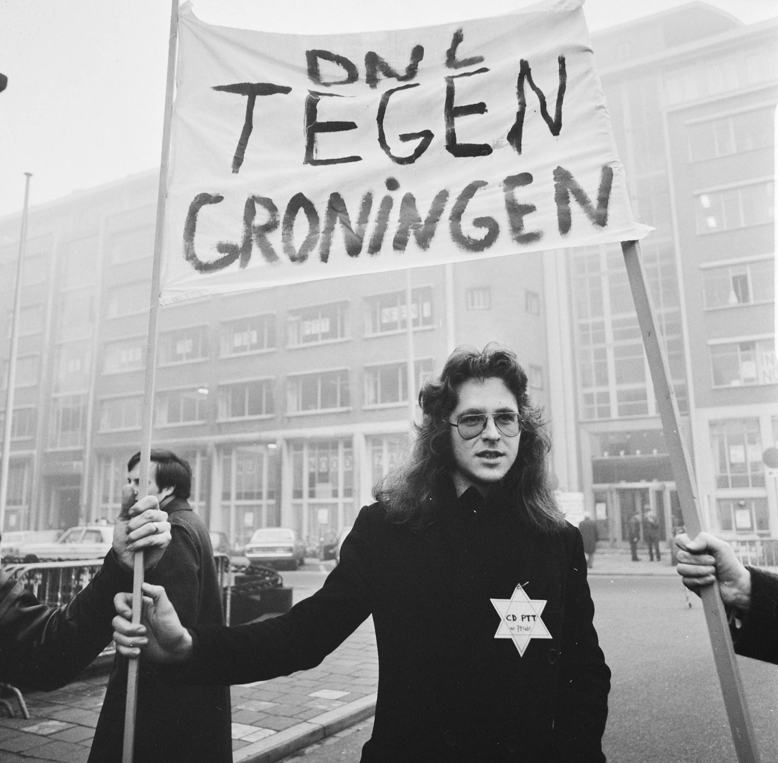 PTT-ers demonstreren in Den Haag voor het kantoor van de PTT i.v.m de voorgenomen verhuizing naar Groningen — Demonstrant met ster op kleding en spandoek — Foto: Mieremet, Rob / Anefo. Licentie: Set B: Rechtenvrij / Publiek Domein. Bron: Nationaal Archief. http://hdl.handle.net/10648/ac4552ea-d0b4-102d-bcf8-003048976d84