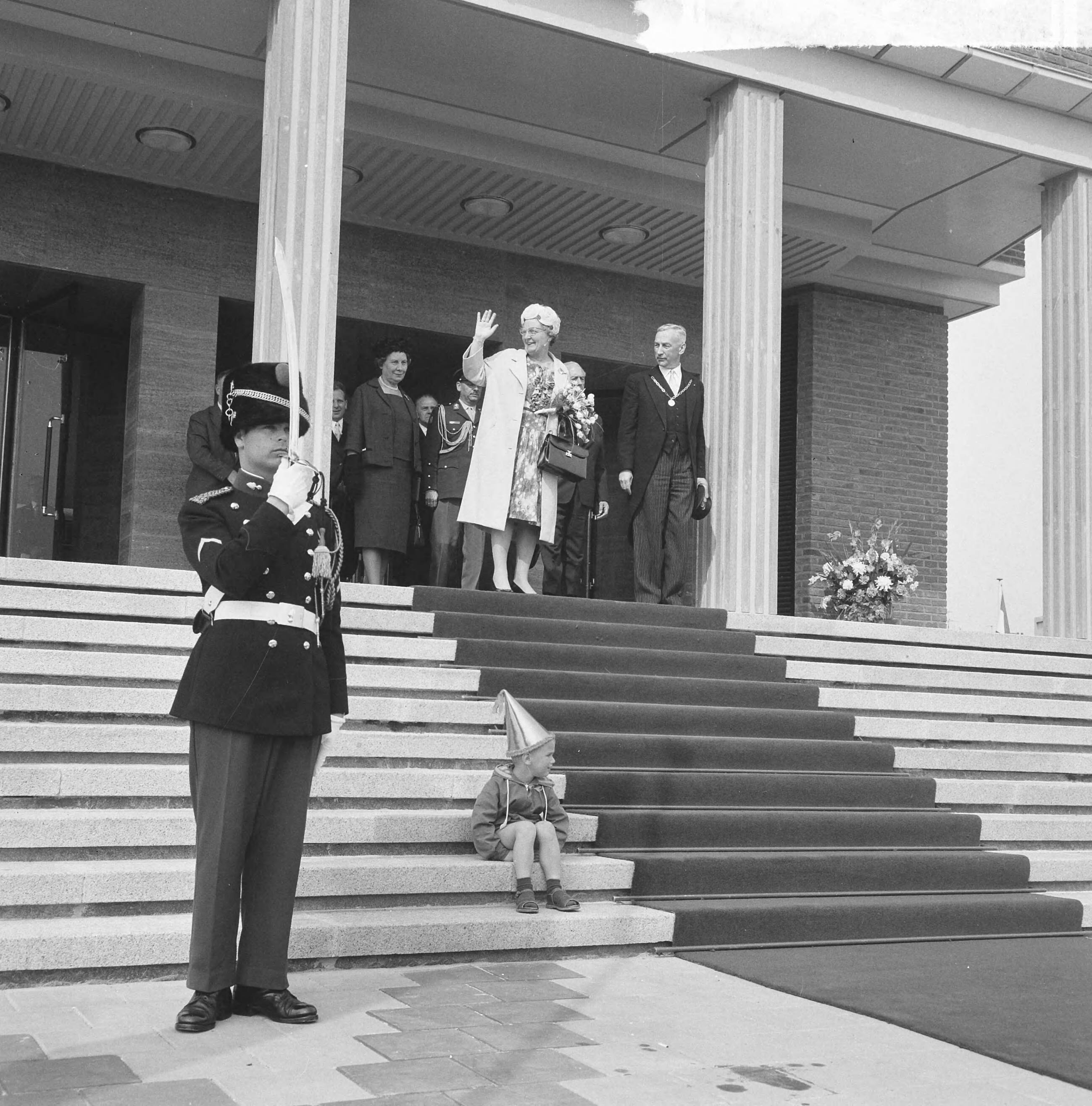 Koningin Juliana opent het nieuwe stadhuis te Vlissingen — Koningin Juliana verlaat het stadhuis, met naast zich burgemeester B. Kolff. Op de trappen zit een jongetje met feestmuts, recht brengt een marechaussee een saluut met zijn sabel — Foto: Nijs, Jac. de / Anefo. Licentie: Set B: Rechtenvrij / Publiek Domein. Bron: Nationaal Archief. http://hdl.handle.net/10648/78b80860-c725-bcde-8d7e-981705b8d5c3