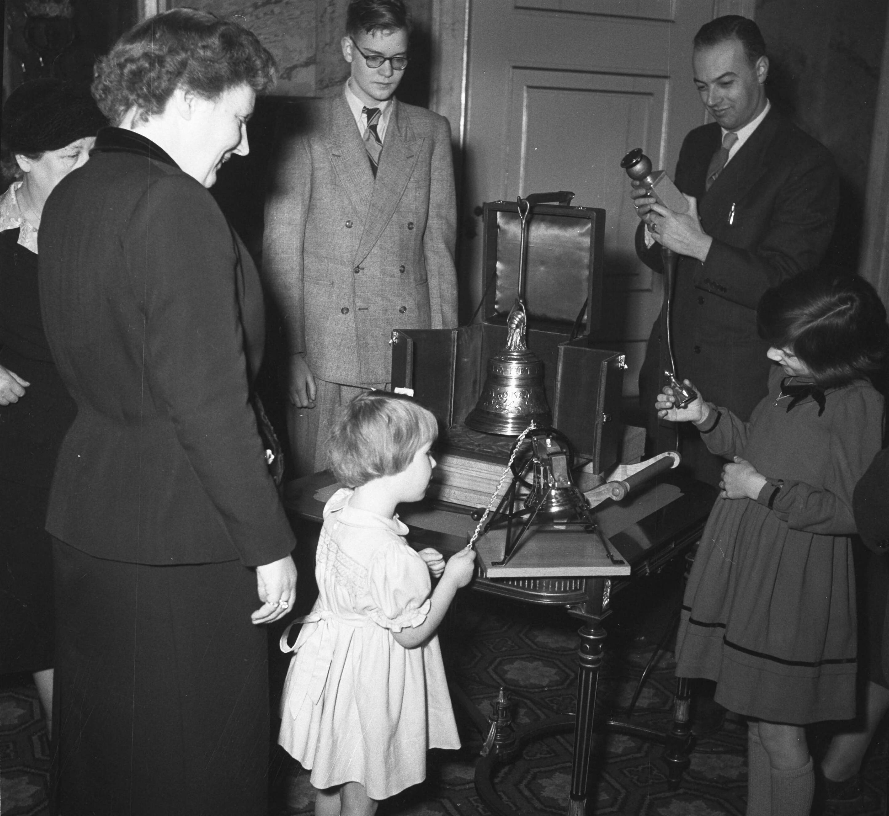 Aanbieding op paleis Soestdijk aan prinses Christina van de kleinste luidklok, het z.g. Marijke-klokje, behorend tot het carillon dat Nederland als dank aan de Verenigde Staten zal geven — Prinses Marijke (Christina) bij het klokje; links koningin Juliana; in het midden Rob Veenstra, rechts Ria Schagen — Foto: Noske, J.D. / Anefo. Licentie: Set B: Rechtenvrij / Publiek Domein. Bron: Nationaal Archief. http://hdl.handle.net/10648/a8f59834-d0b4-102d-bcf8-003048976d84
