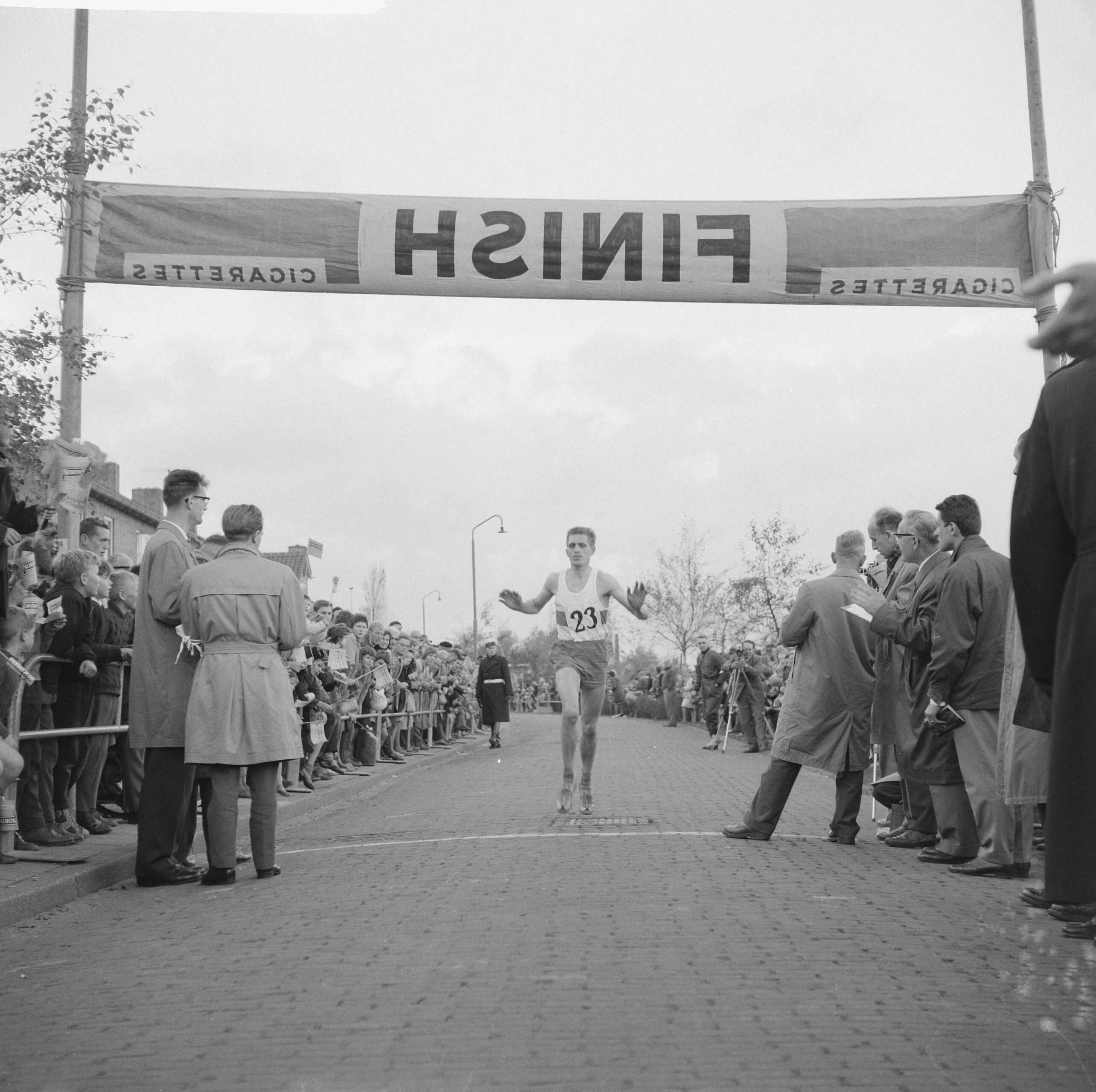 Fotocollectie Anefo — Marathonloop in Zaandam [een atleet (met startnummer 23) gaat over de finish] — Foto: Brinkman, Dave / Anefo. Licentie: Set B: Rechtenvrij / Publiek Domein. Bron: Nationaal Archief. http://hdl.handle.net/10648/a9e9bc48-d0b4-102d-bcf8-003048976d84