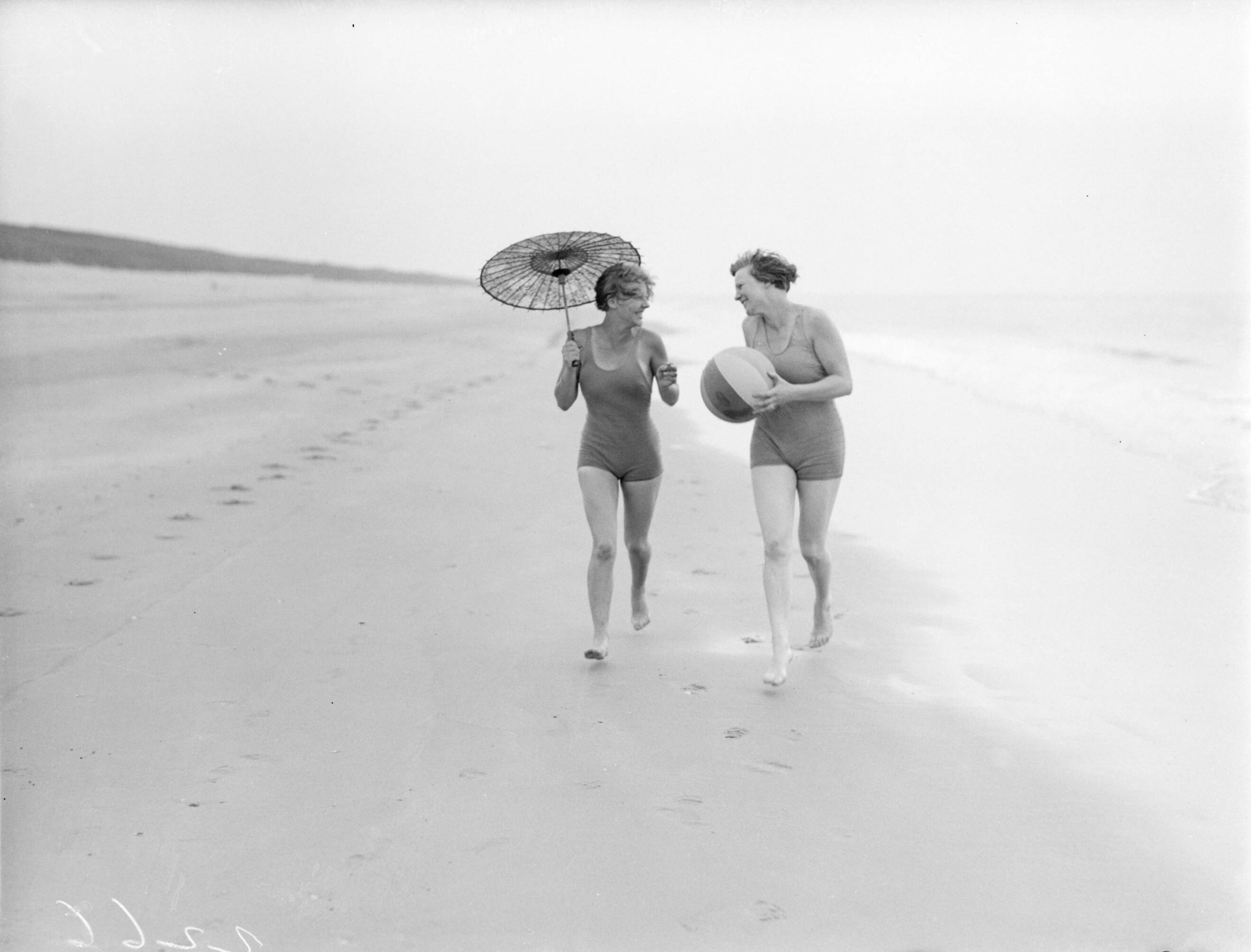 Reportage fotomodel — Model Eva Waldschmidt (met parasol) en Van de Polls vrouw Nell Langlais lopen langs de vloedlijn in Zandvoort — Foto: Poll, Willem van de. Licentie: Set B: Rechtenvrij / Publiek Domein. Bron: Nationaal Archief. http://hdl.handle.net/10648/aea8000a-d0b4-102d-bcf8-003048976d84
