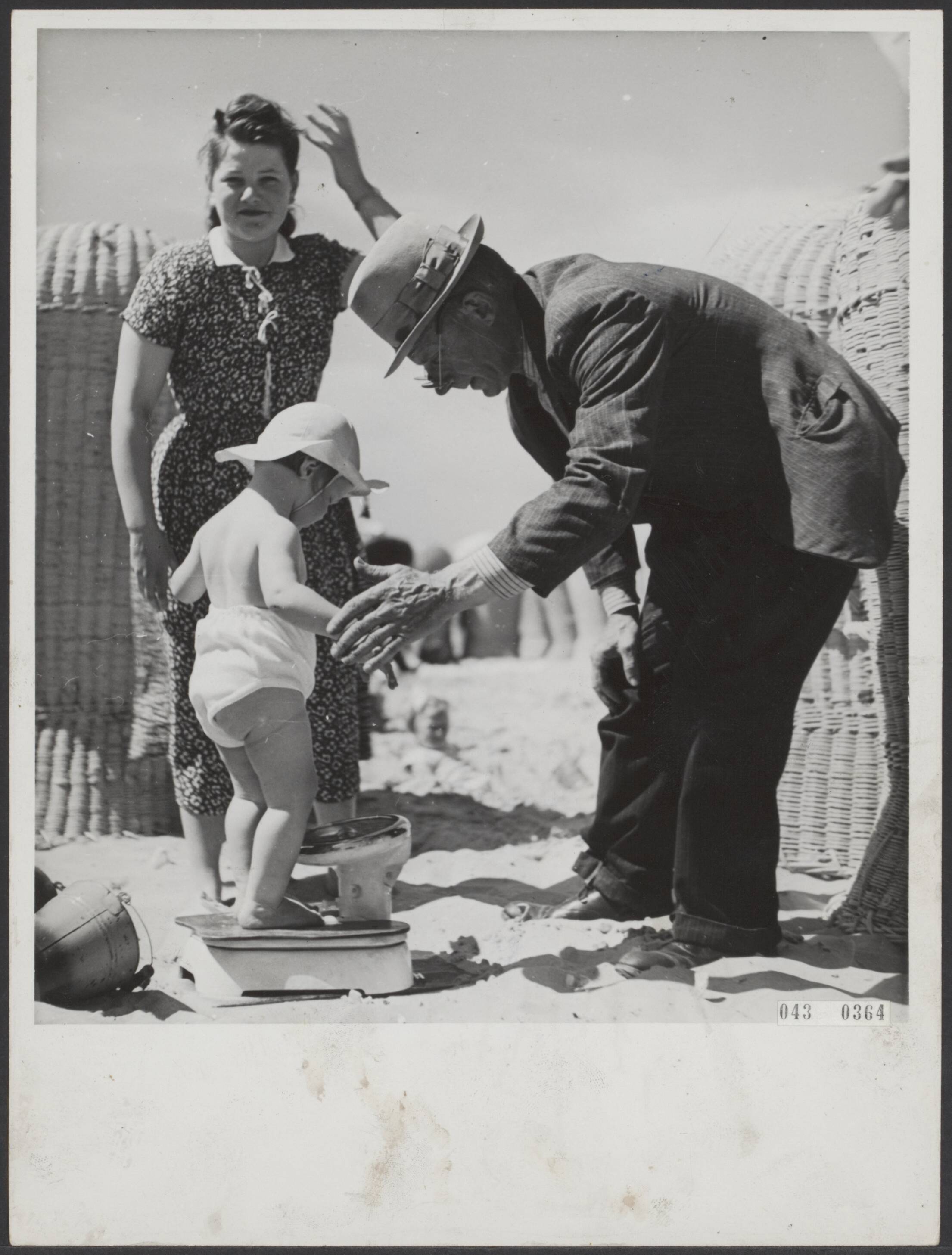 Fotocollectie Elsevier Binnenland — [Serie "Summum". over vermaak in Scheveningen] Nr. 13. Man met weegschaal gaat rond over het strand van Scheveningen. Men kan zich laten wegen tegen betaling (5 cent). Leentje is 18 maanden oud en weegt precies 11 kilo — Foto: Associated Correspondents. Licentie: Set B: Rechtenvrij / Publiek Domein. Bron: Nationaal Archief. http://hdl.handle.net/10648/adf8c892-d0b4-102d-bcf8-003048976d84