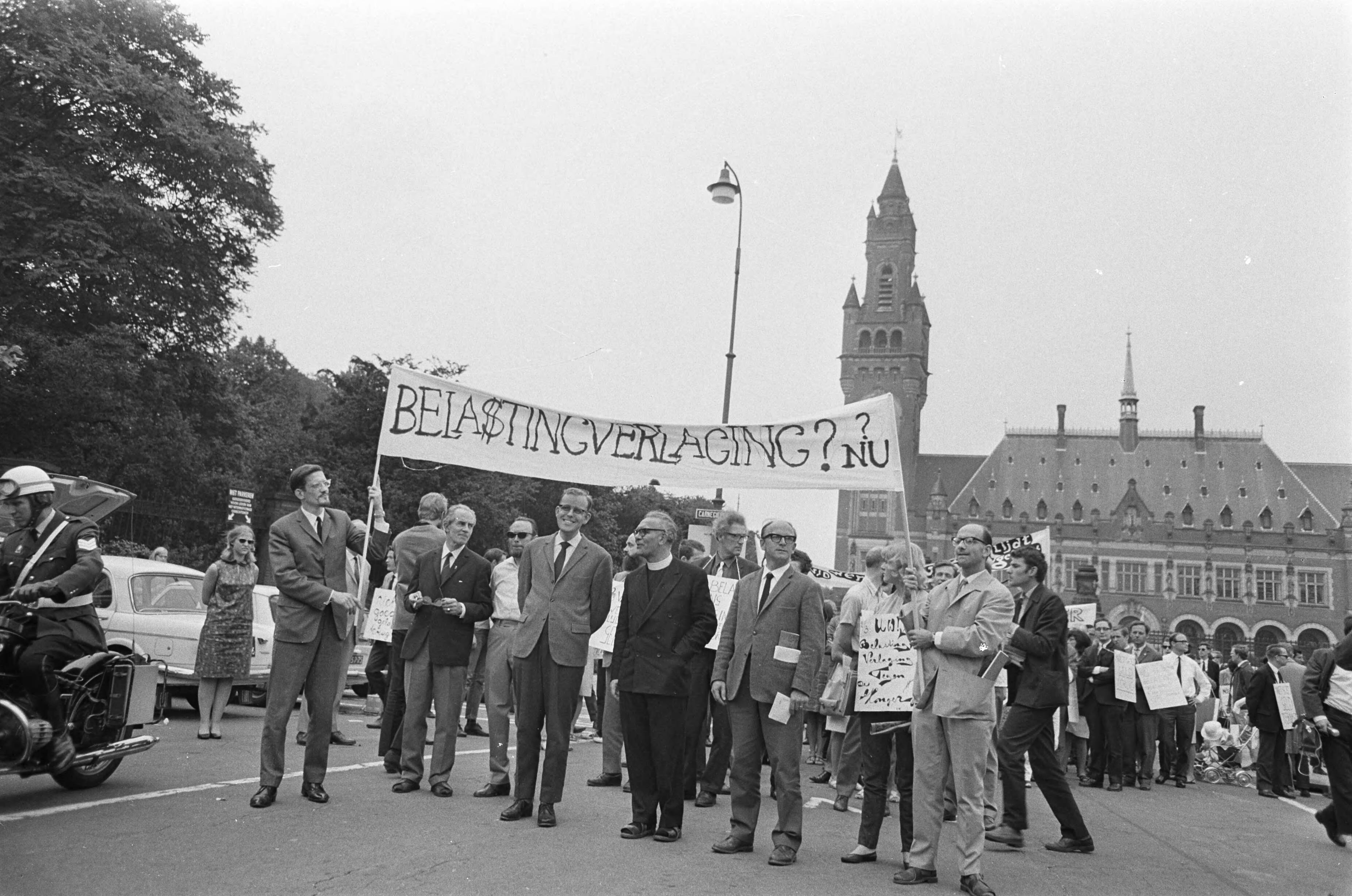 Sjaloomgroep brengt geld terug aan het rijk — Met spandoeken vanaf het Vredespaleis te Den Haag met enveloppe in de hand — Foto: Fotograaf Onbekend / Anefo. Licentie: Set B: Rechtenvrij / Publiek Domein. Bron: Nationaal Archief. http://hdl.handle.net/10648/ab1dc136-d0b4-102d-bcf8-003048976d84
