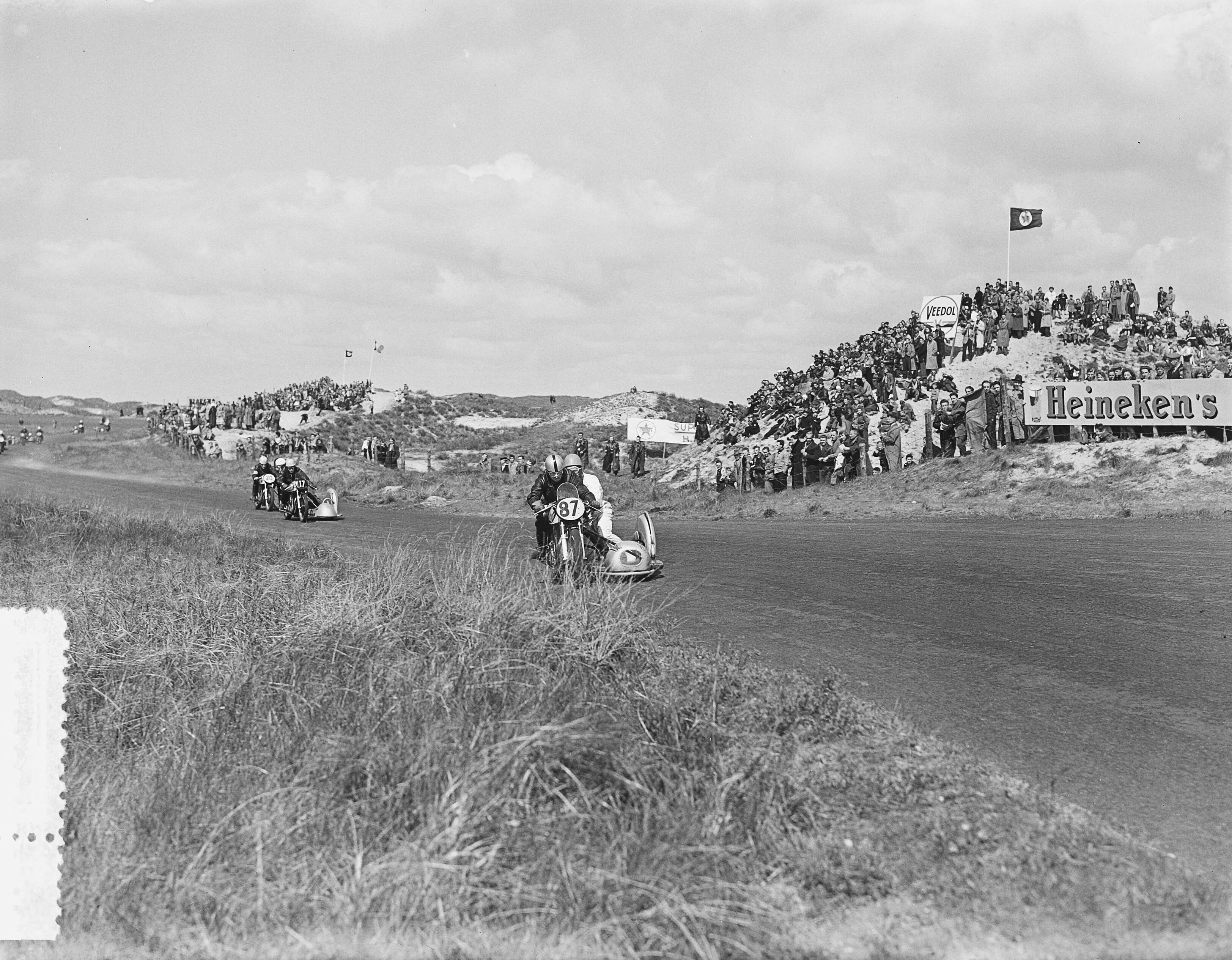 Fotocollectie Anefo — Motorraces Zandvoort. Steeman (87) gevolgd door (17) Wijns( B) winnaar met publiek — Foto: Bilsen, Joop van / Anefo. Licentie: Set B: Rechtenvrij / Publiek Domein. Bron: Nationaal Archief. http://hdl.handle.net/10648/a90bc30c-d0b4-102d-bcf8-003048976d84