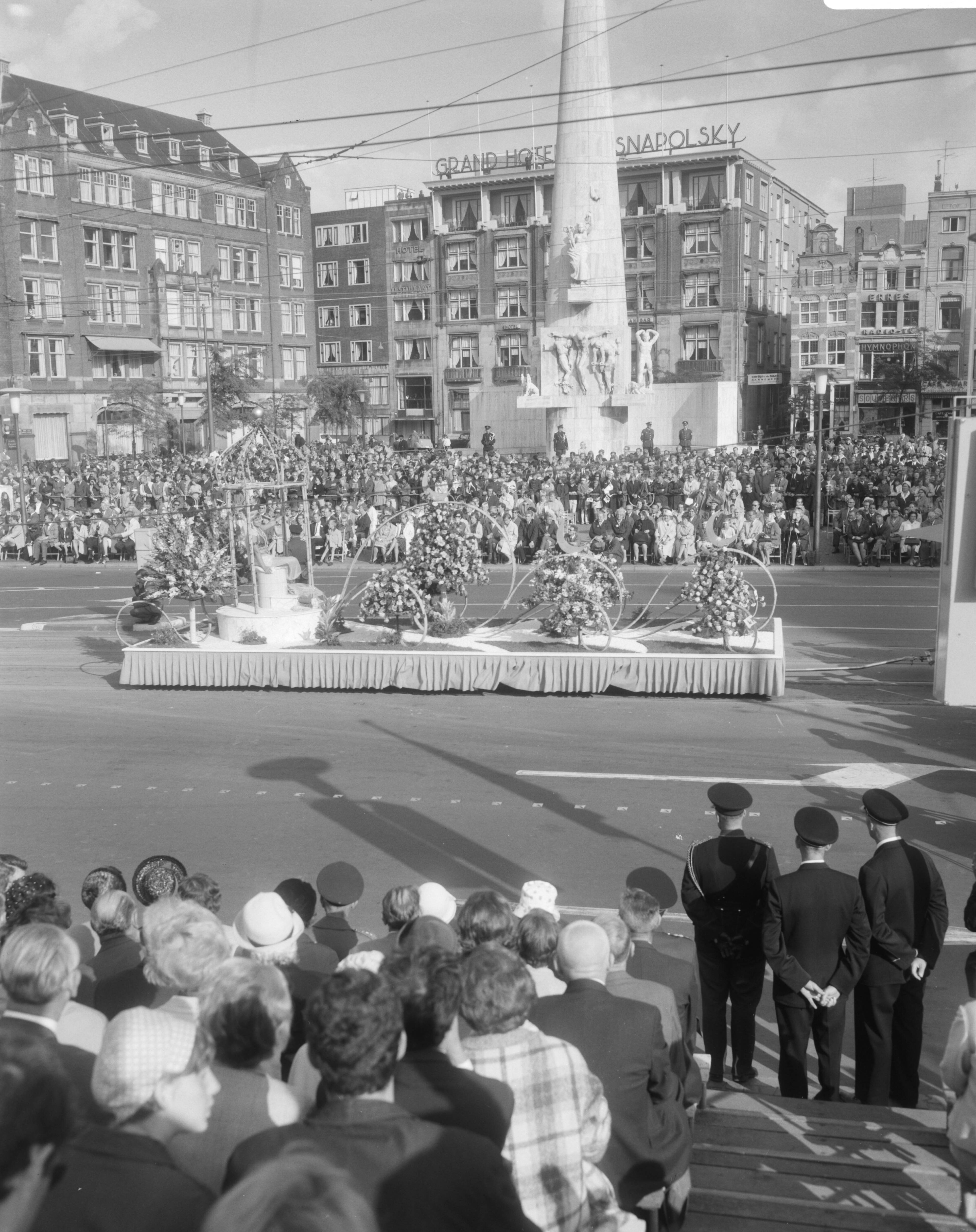 Bloemencorso van Aalsmeer te Amsterdam — Een praalwagen bij de Dam te Amsterdam met op de achtergrond het Nationaal Monument — Foto: Bilsen, Joop van / Anefo. Licentie: Set B: Rechtenvrij / Publiek Domein. Bron: Nationaal Archief. http://hdl.handle.net/10648/aafaf3f4-d0b4-102d-bcf8-003048976d84