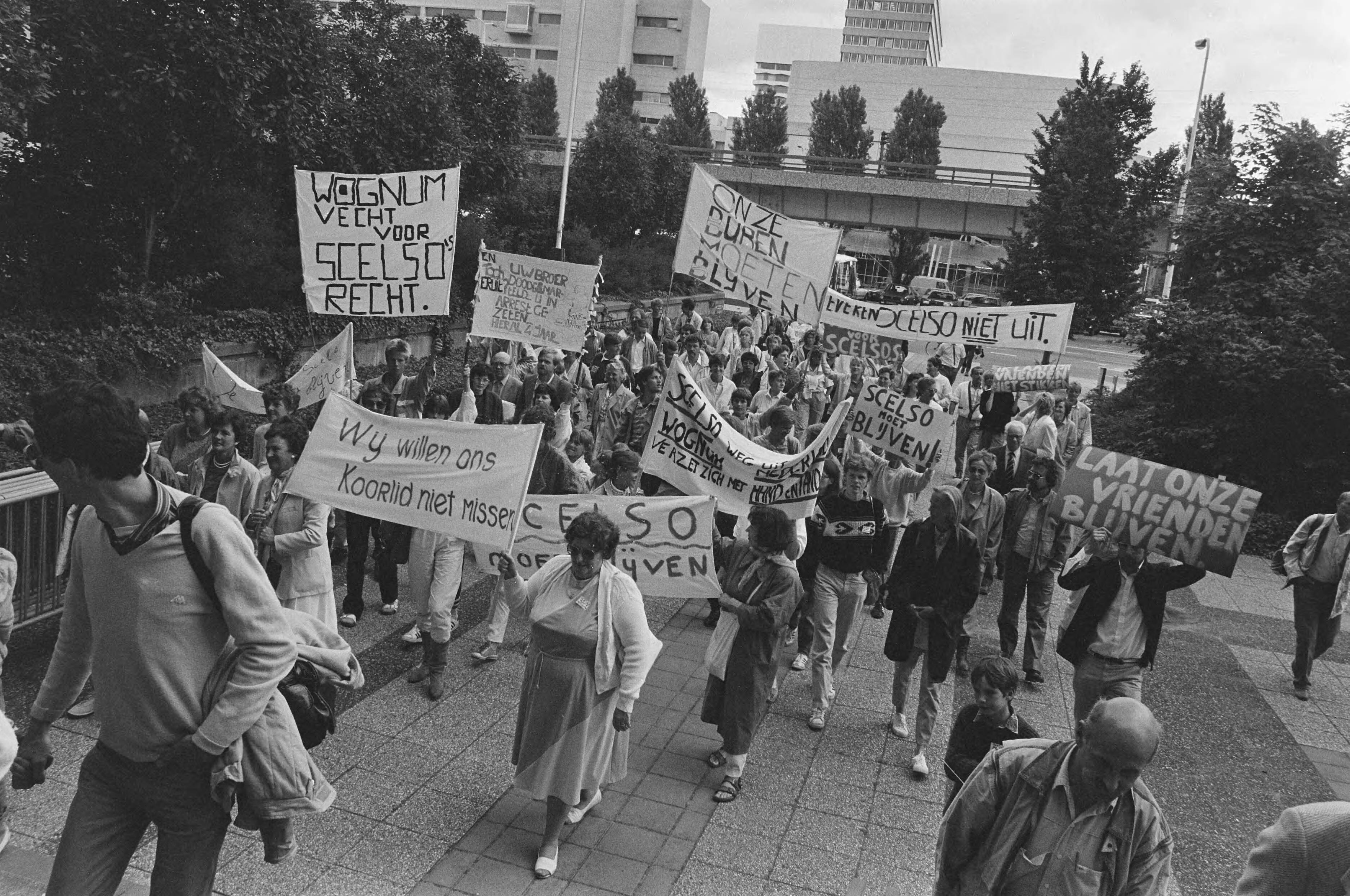 Fotocollectie Anefo — Kort geding inwoners Wognum tegen uitwijzing Argentijns gezin Scelso; aankomst inwoners met spandoeken bij Paleis van Justitie Den Haag — Foto: Bogaerts, Rob / Anefo. Licentie: Set B: Rechtenvrij / Publiek Domein. Bron: Nationaal Archief. http://hdl.handle.net/10648/ad486ed4-d0b4-102d-bcf8-003048976d84